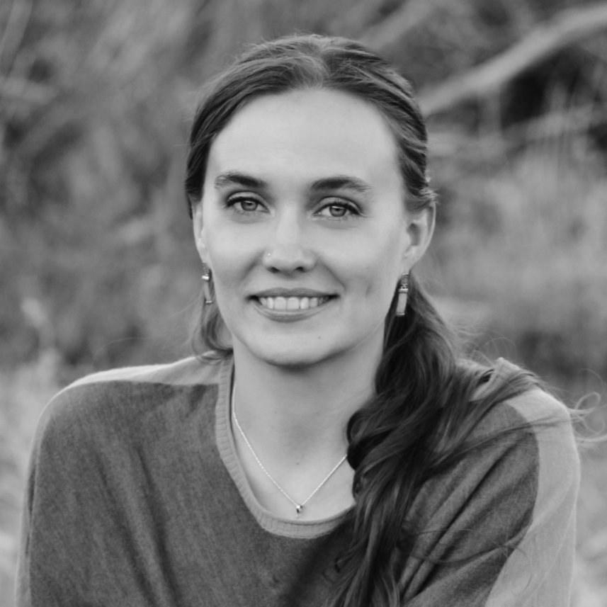 A black and white portrait of a smiling woman with long hair, wearing a sweater and jewelry, outdoors.