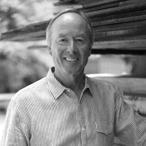 A smiling middle-aged man in a striped button-up shirt standing outdoors near wooden logs.