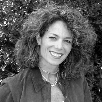 Black and white portrait of a woman with curly hair smiling, outdoors with foliage in the background.