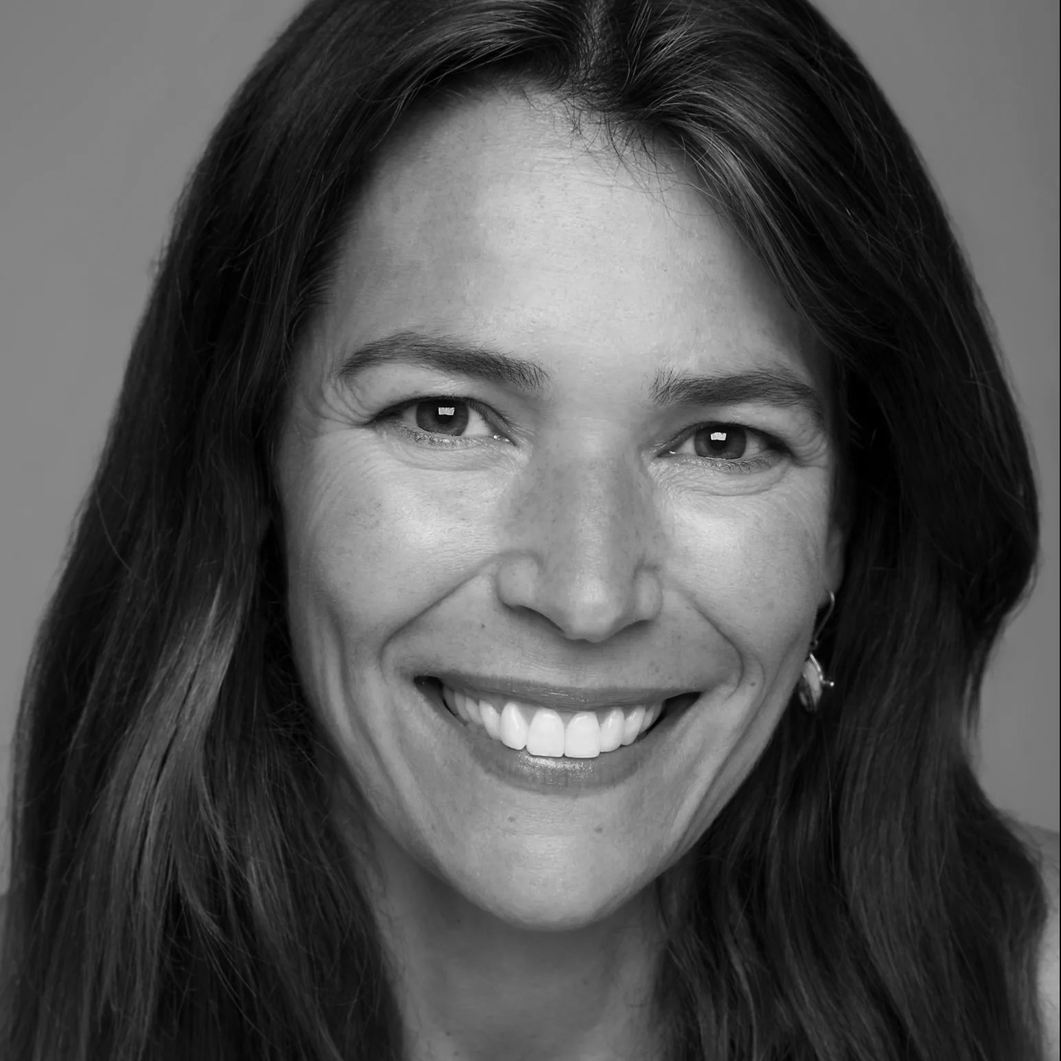 Black and white close-up portrait of a woman with long dark hair, smiling, showing her teeth, with earrings, against a plain background.
