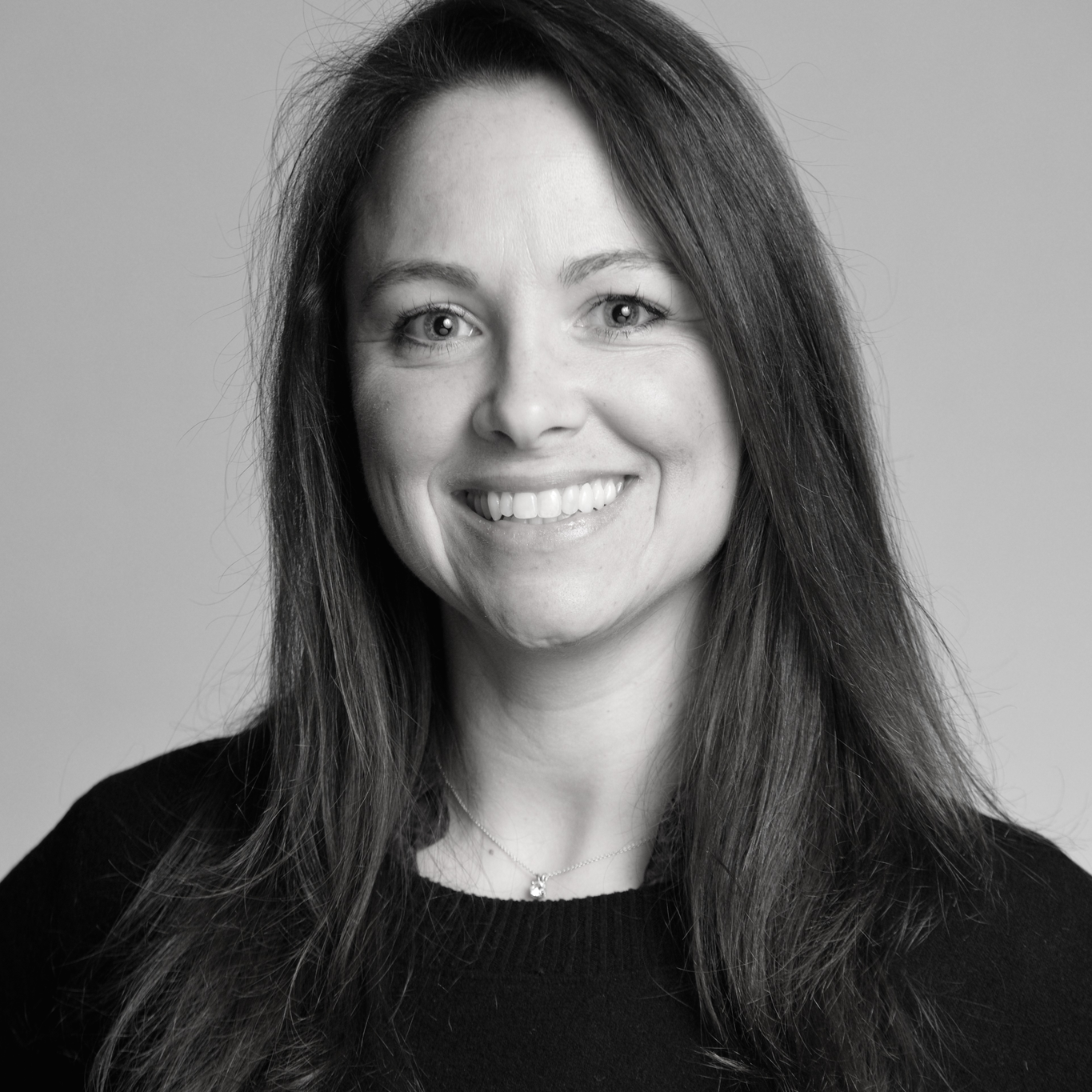 Black and white portrait of a woman with long, dark hair, smiling and wearing a dark top and a necklace.