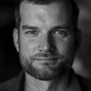 Close-up black-and-white portrait of a man with short hair and a beard, looking directly at the camera with a neutral expression.