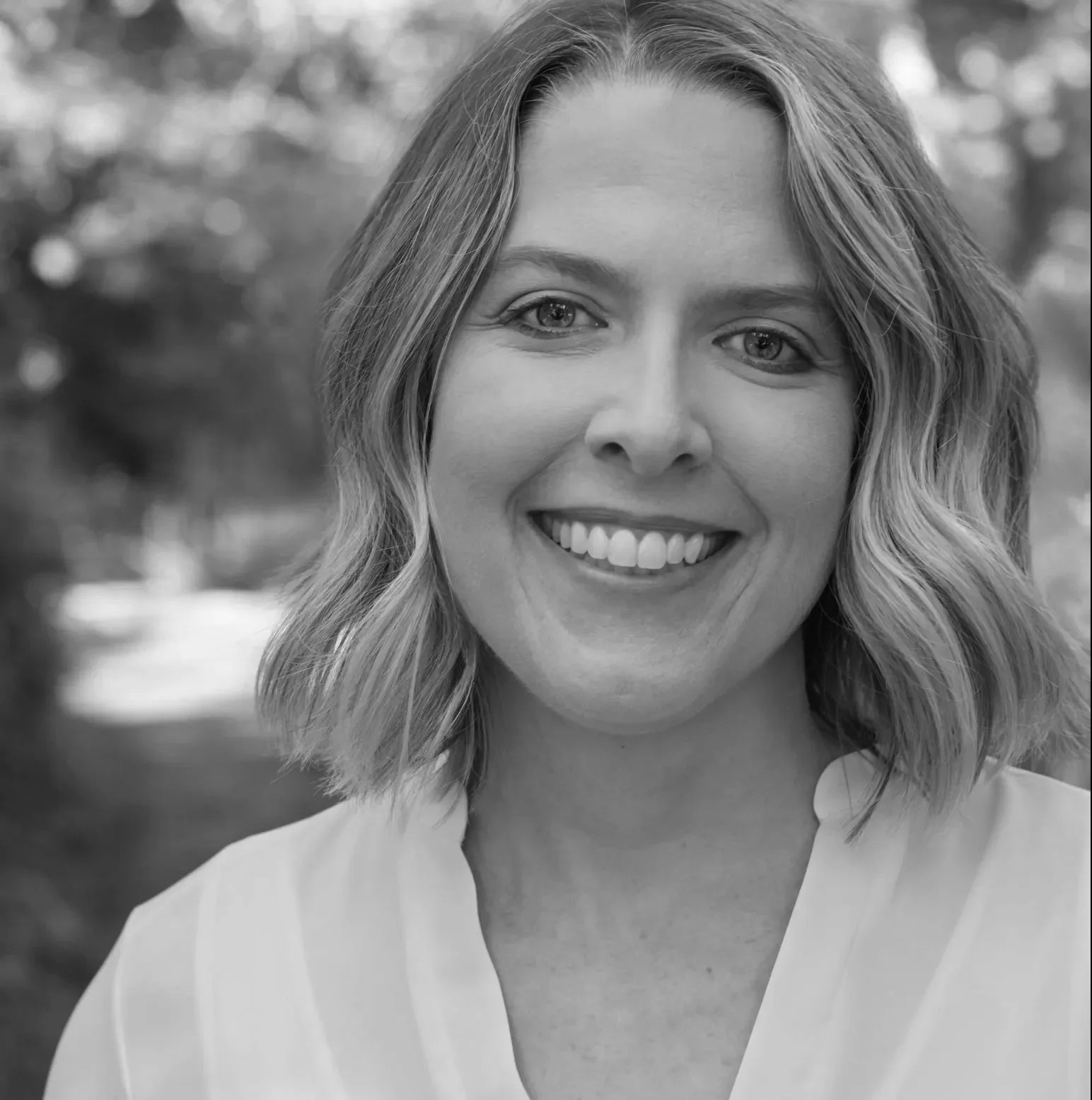 Black and white portrait of a smiling woman with shoulder-length wavy hair outdoors.