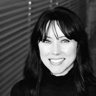 Black and white photo of a woman with long dark hair, smiling, wearing a black turtleneck, in front of a background with horizontal blinds.
