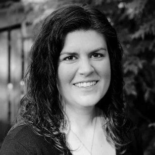 Black and white portrait of a smiling woman with wavy dark hair outside, blurred foliage background.