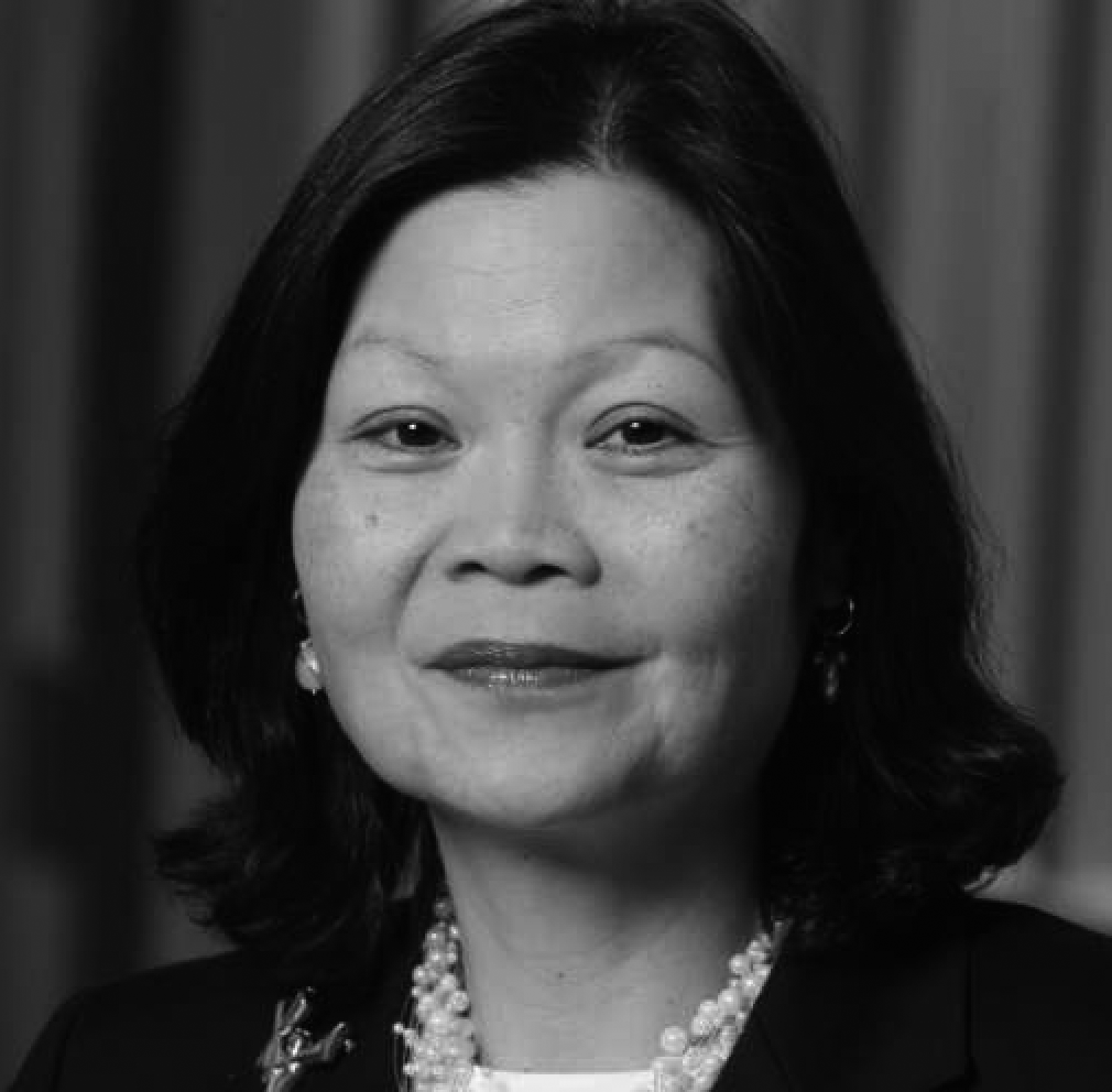 Black and white portrait of a woman with shoulder-length hair, wearing earrings, a necklace with multiple strands, and a dark blazer, smiling softly.