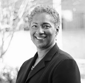 A portrait of a woman with short curly hair, wearing a blazer, smiling outdoors.