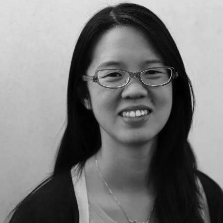 Black and white photo of a young woman with long dark hair, wearing glasses, a necklace, and a light-colored top, smiling gently.
