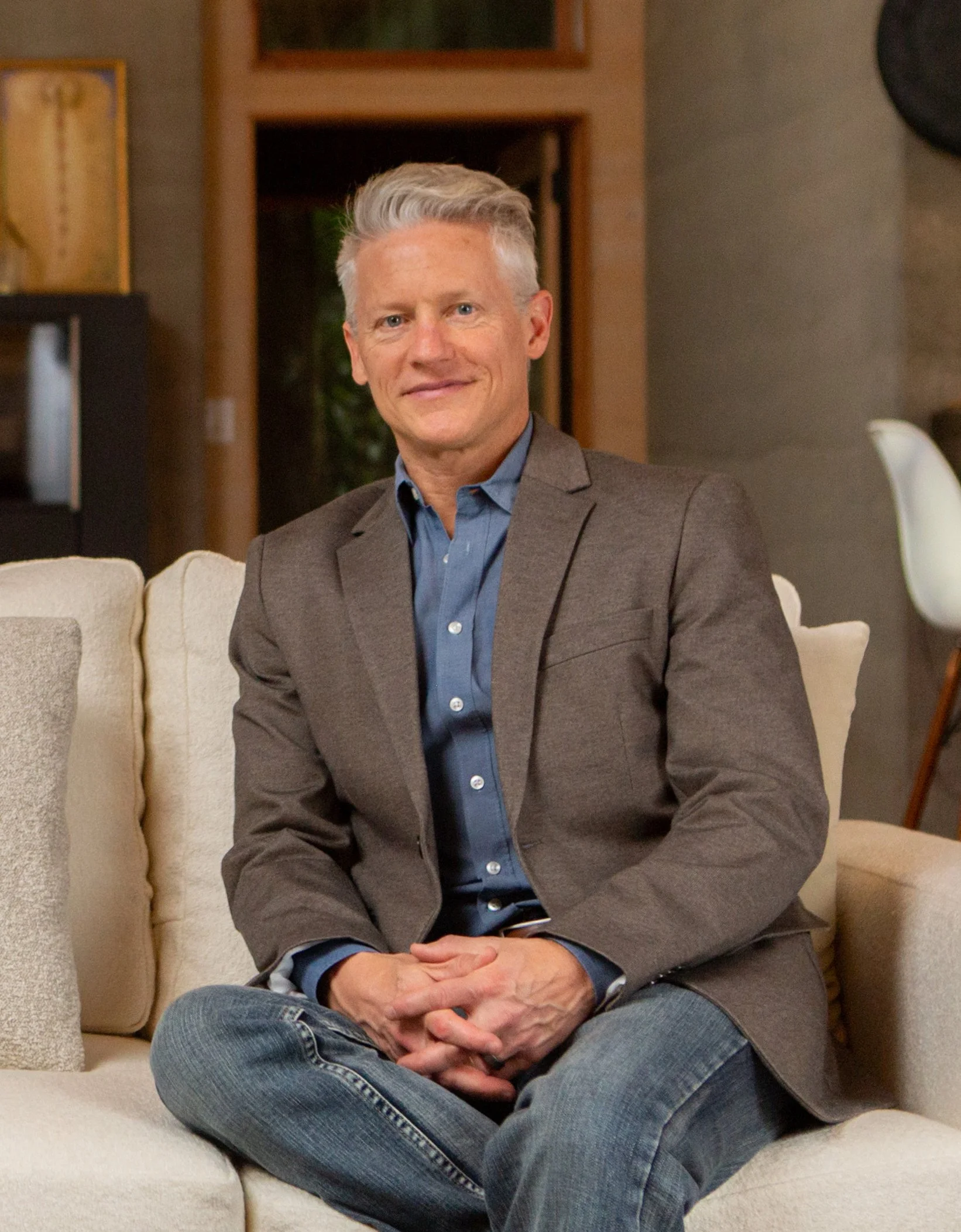 A middle-aged man with gray hair, dressed in a brown blazer and blue shirt, sitting on a beige couch in a cozy living room.