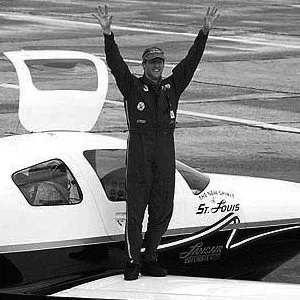 Person in racing suit standing with arms raised next to a boat with the name St. Louis.