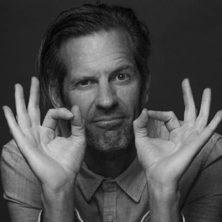Man with slightly long hair and beard making a gesture with both hands near his face in black and white