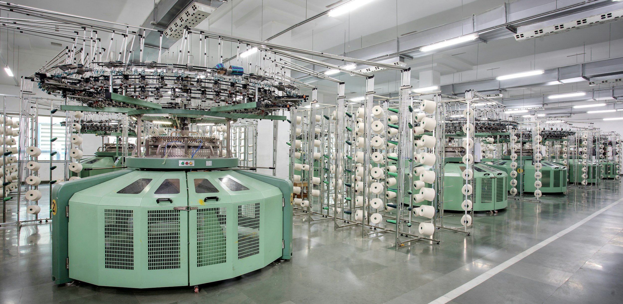 Industrial textile manufacturing facility with large circular knitting machines surrounded by racks of white and green yarn spools.
