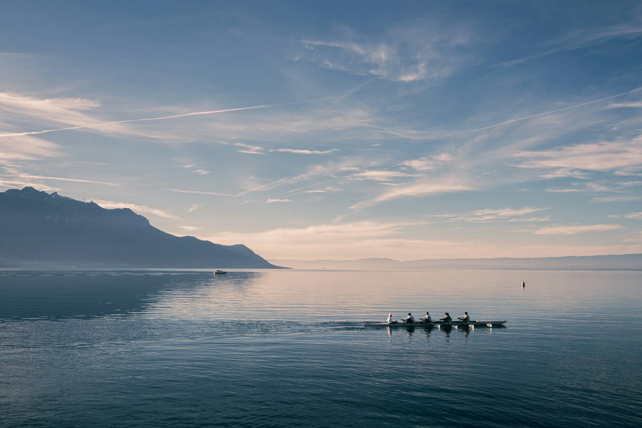 A peaceful lake scene with a mountain in the background, a small boat in the distance, and a rowing team in a boat in the foreground on calm water under a blue sky with scattered clouds.