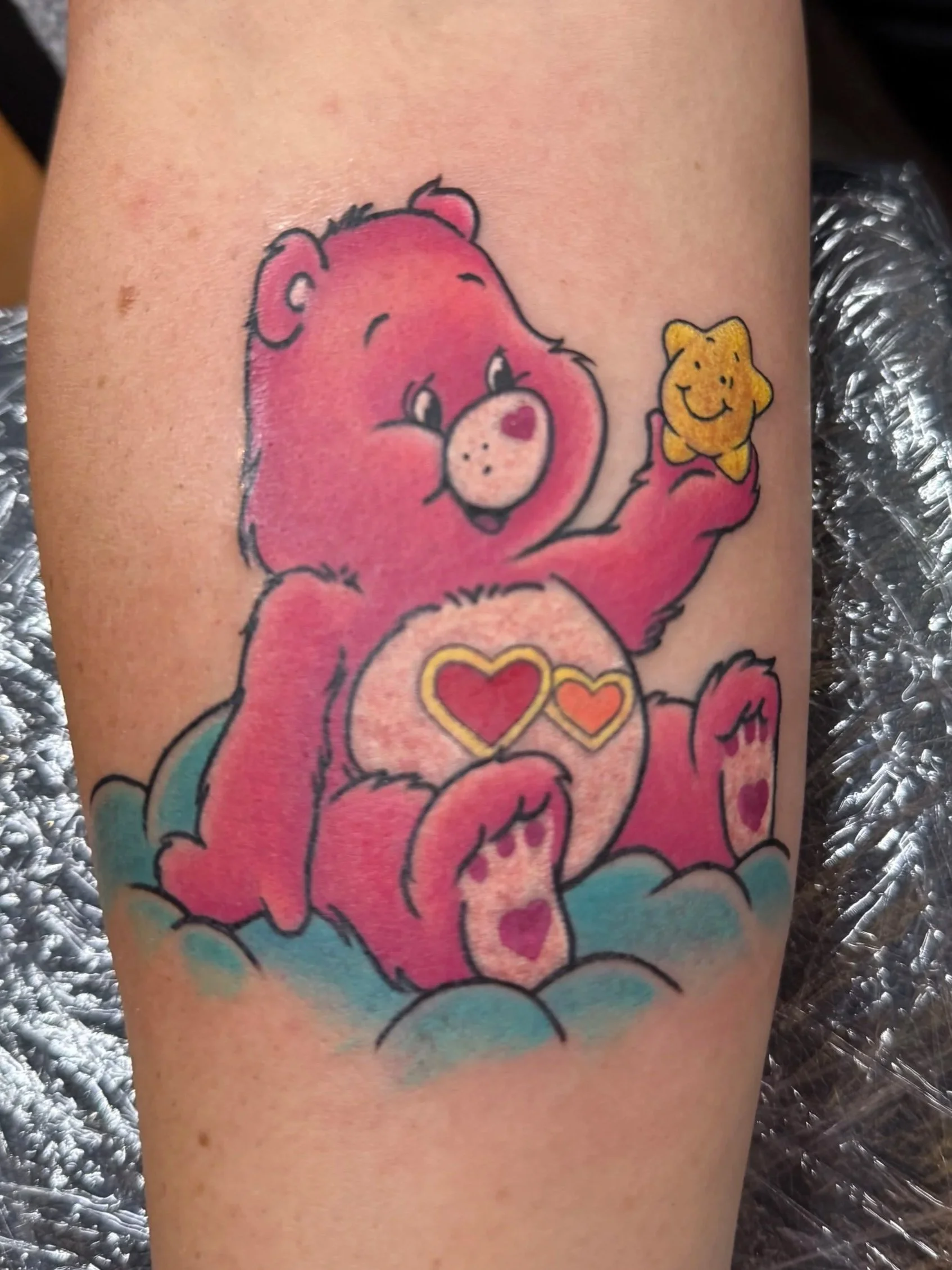 Colorful tattoo of a pink teddy bear holding a small yellow star with a smiley face. The bear is sitting on clouds and has hearts on its paws and chest, with some hearts depicted in the bear's eyes.