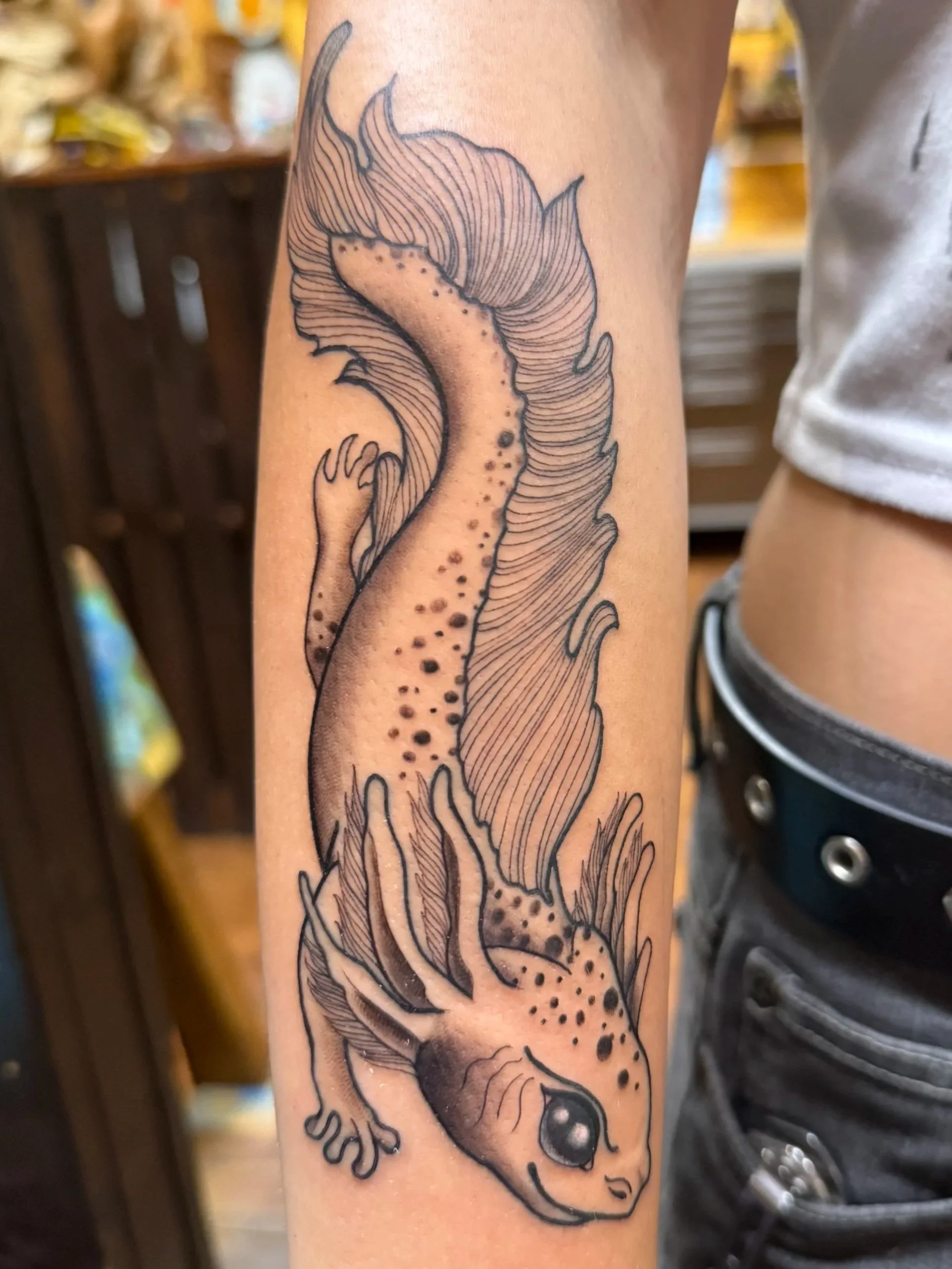 Black and gray tattoo of a salamander or newt on a person's forearm, with detailed scales, limbs, and a large eye.
