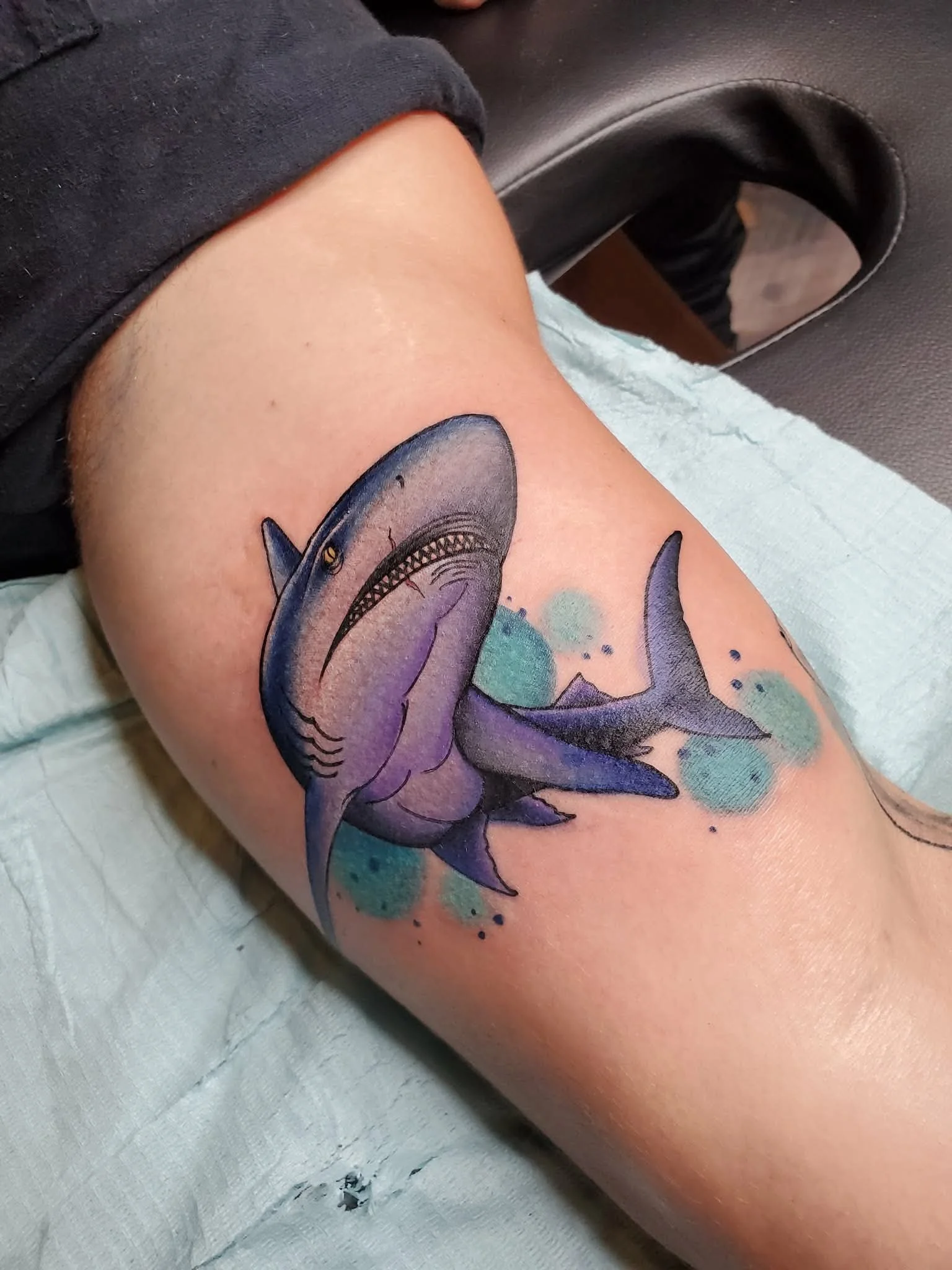 Tattoo of a shark on a person's upper arm, with purple and blue watercolor-like background effects.