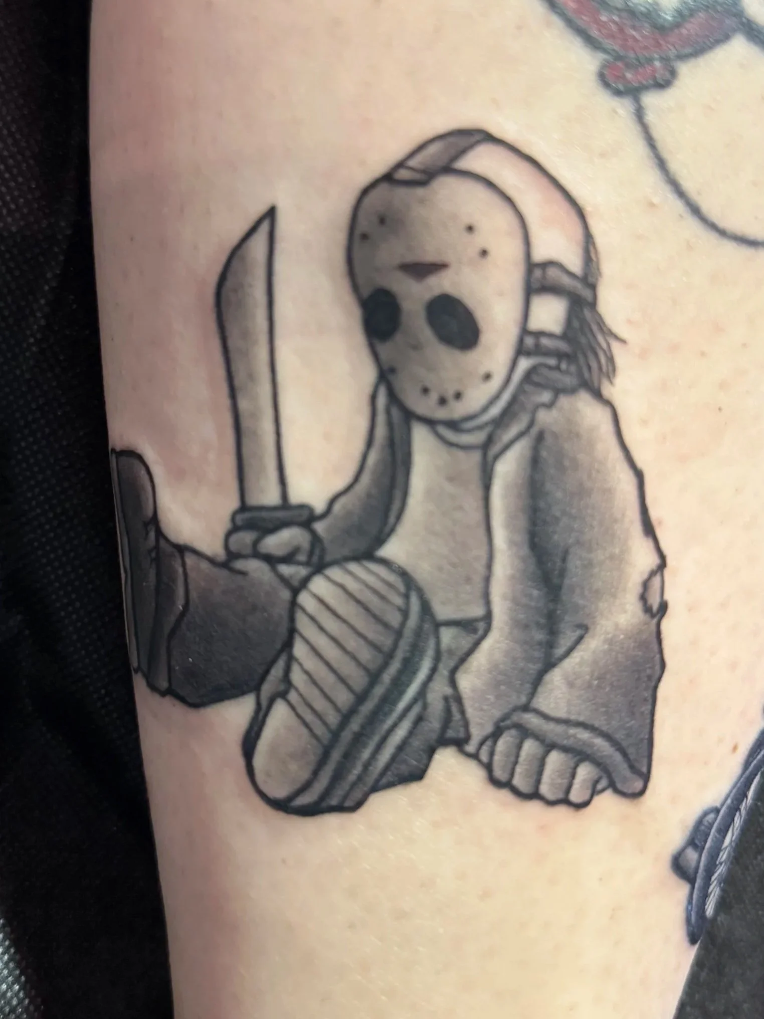 Tattoo of a horror character Jason Vorhees wearing a hockey mask, hoodie, and sneakers, sitting on the ground.