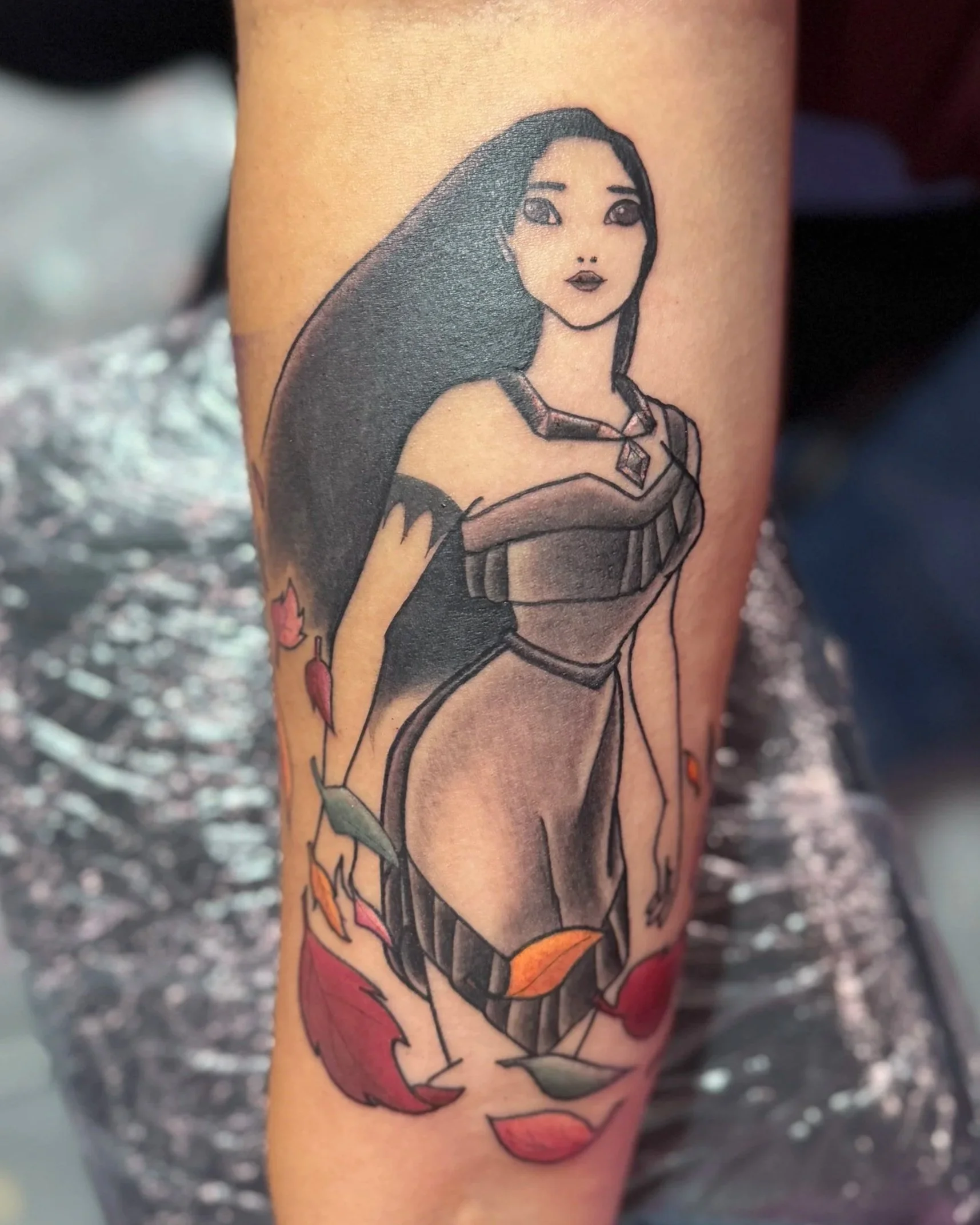 Tattoo of a Disney character Pocahontas with long black hair, wearing a black and gray dress, surrounded by falling colorful autumn leaves.
