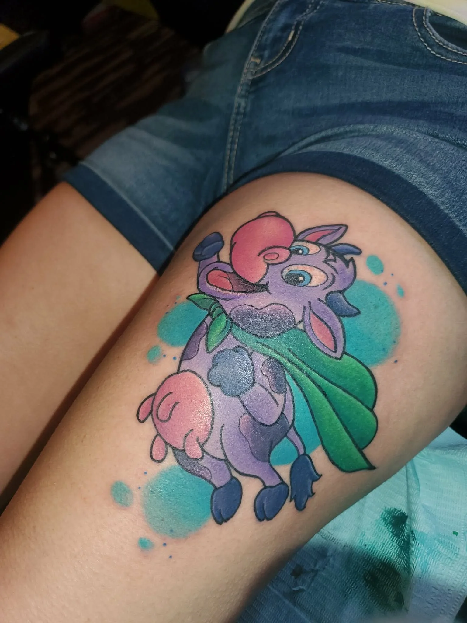 Colorful tattoo of a cartoon cow with pink udders, large blue eyes, and a mischievous expression, surrounded by aqua blue and teal splashes, on a person's thigh.