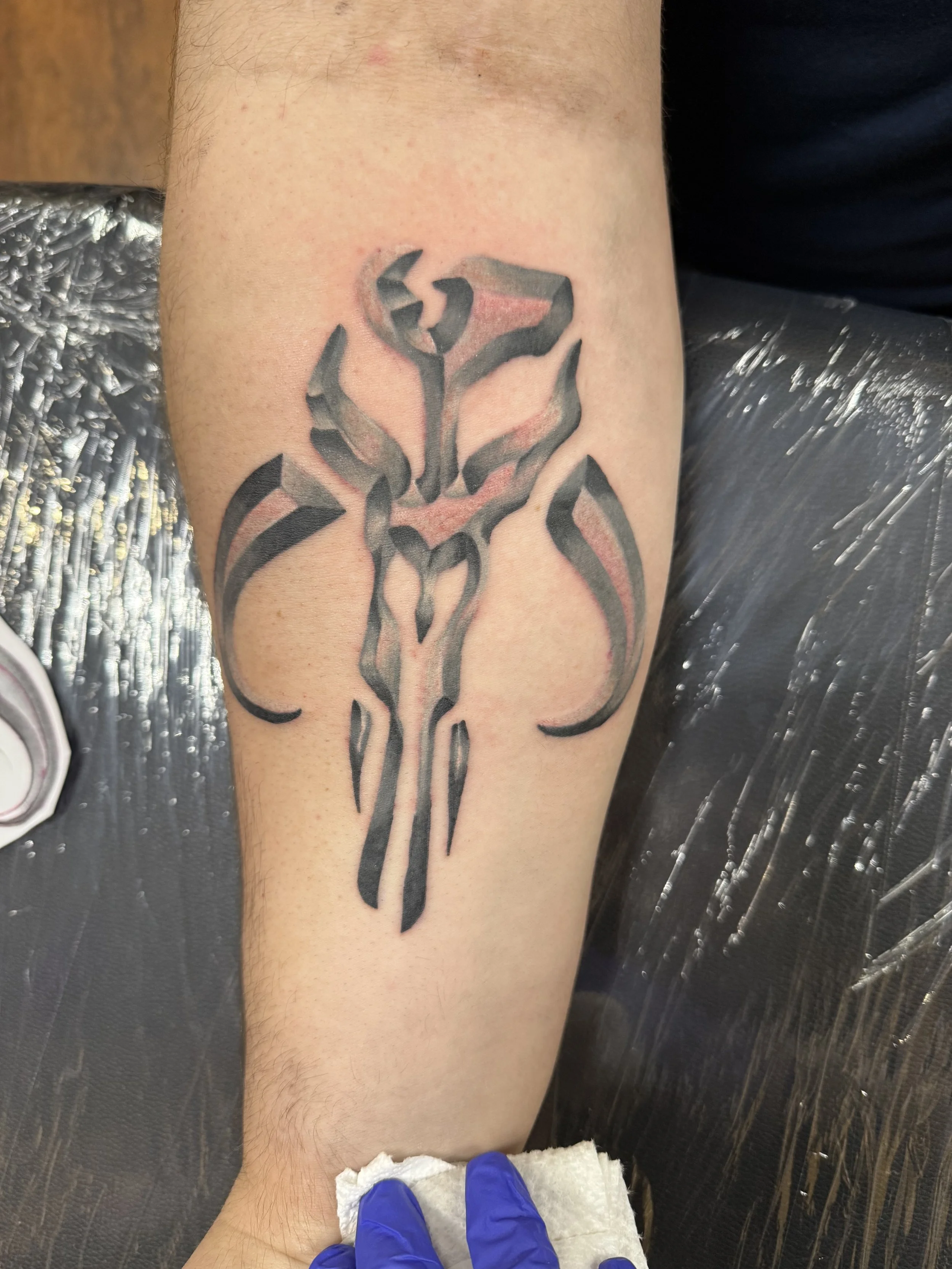 Black tribal-style tattoo of a stylized Star Wars Mandalorian symbol on a person's forearm.