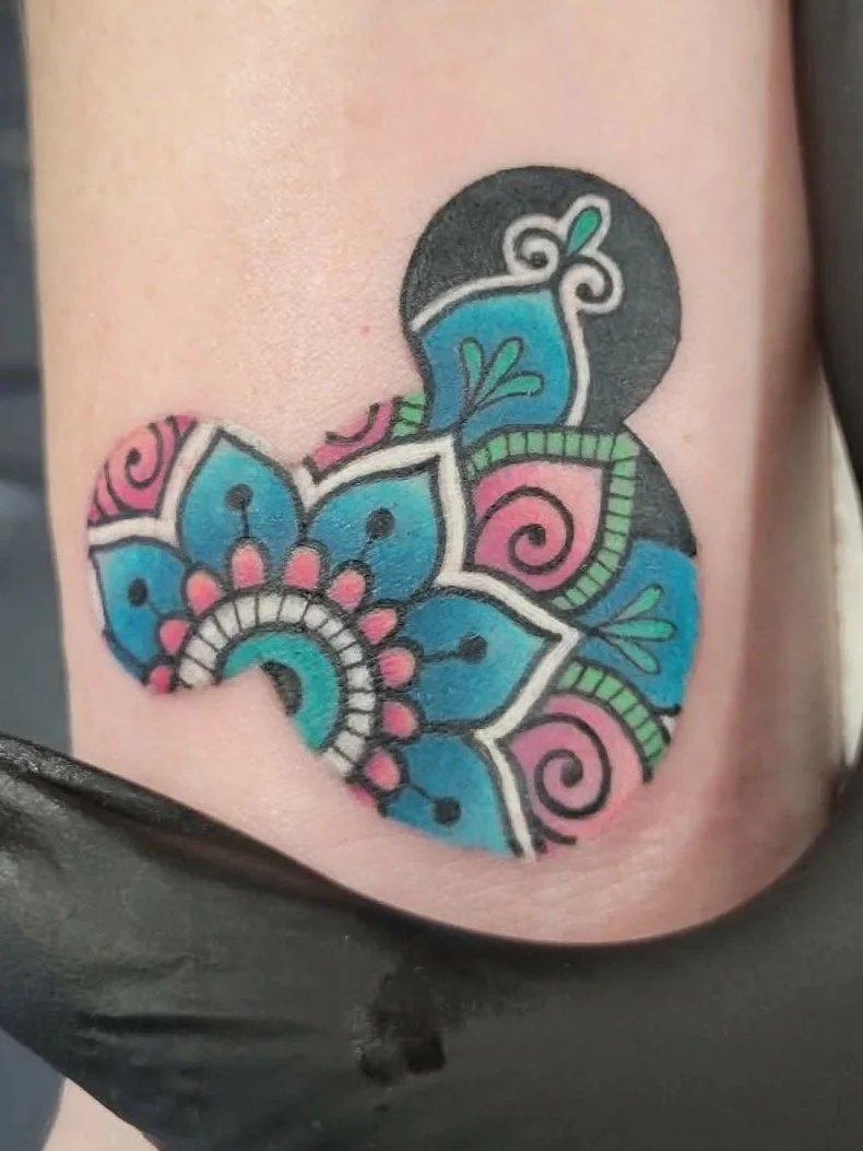 Colorful tattoo of a floral and abstract pattern with pink, blue, green, and black ink on skin.