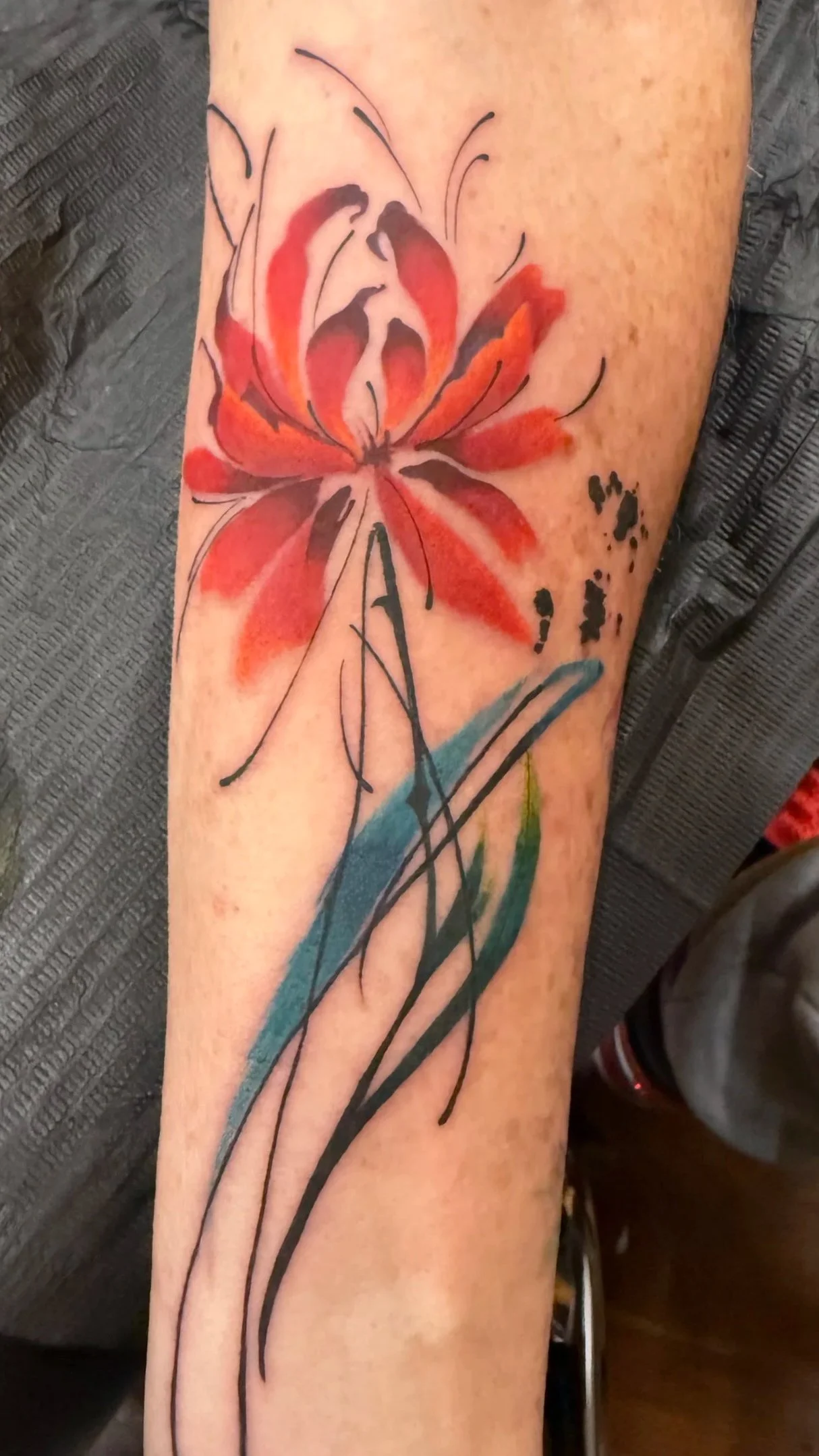 Tattoo of a red and orange flower with black and teal abstract lines on a person's arm.