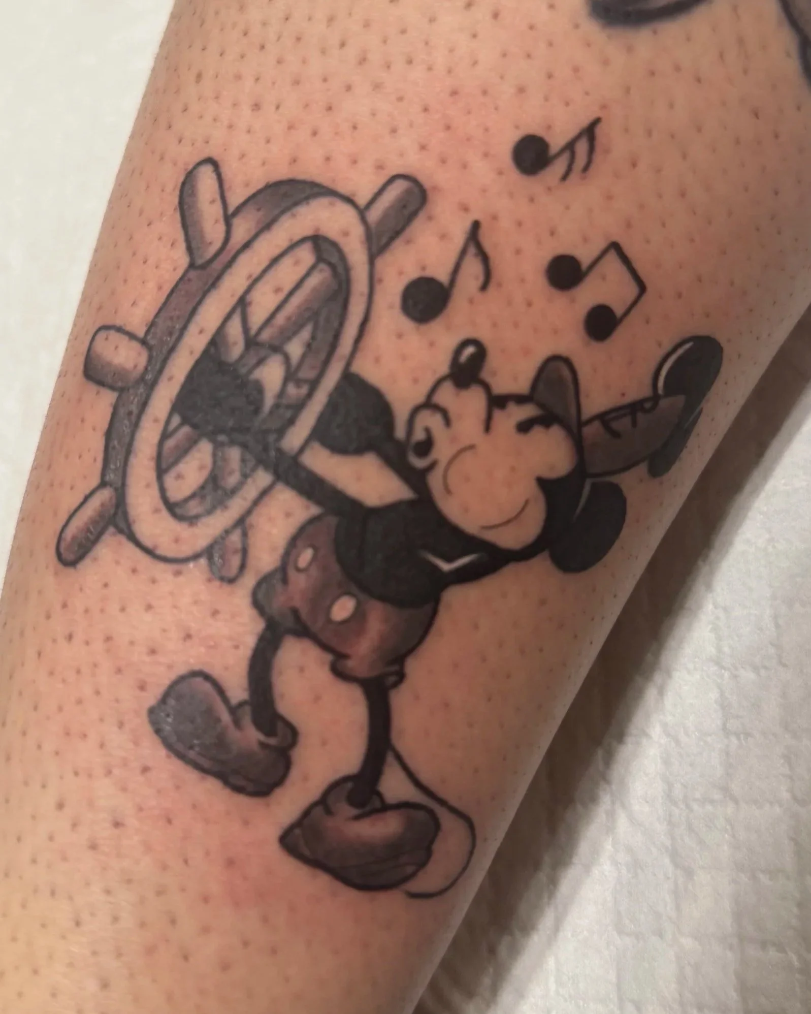 Tattoo of Mickey Mouse carrying a ship's steering wheel, with musical notes around.