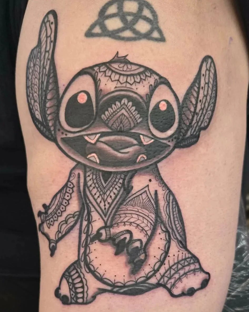 Black tattoo of a stylized cartoon Disney Stitch with large eyes, decorated with tribal patterns and designs.