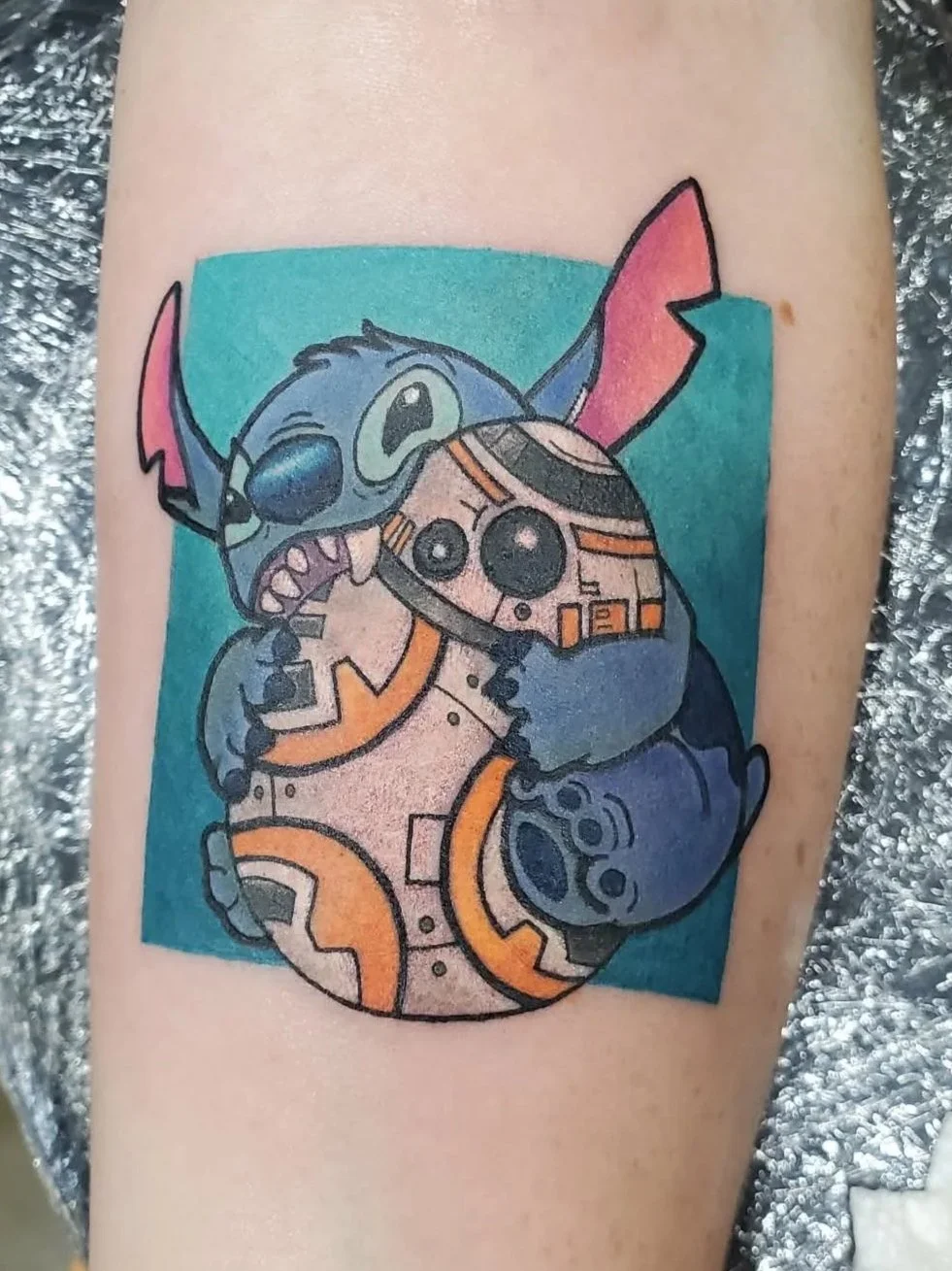 A colorful tattoo of Stitch from Disney's "Lilo & Stitch" holding a small BB-8 droid from "Star Wars," set against a teal background.