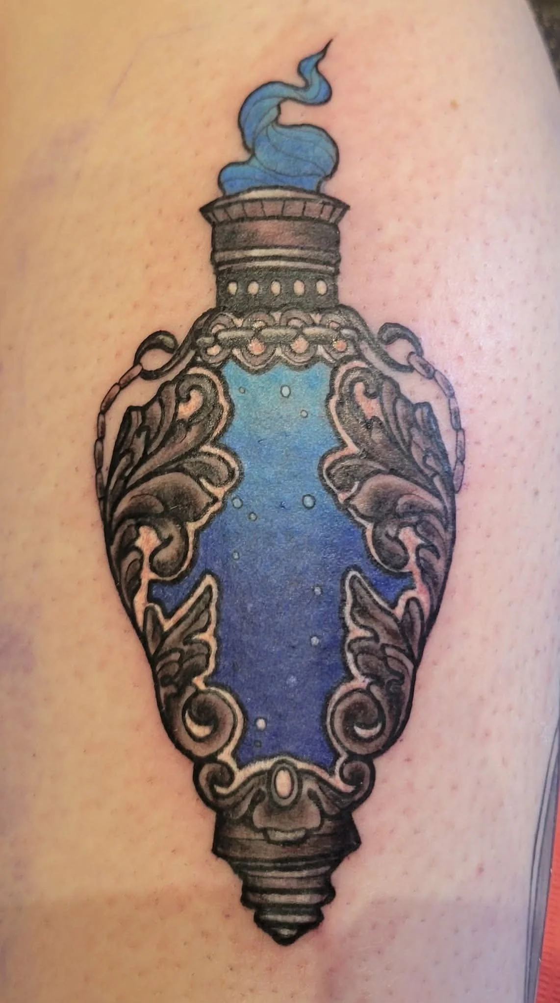 Tattoo of an ornate perfume bottle with swirling patterns and blue liquid inside, on human skin.