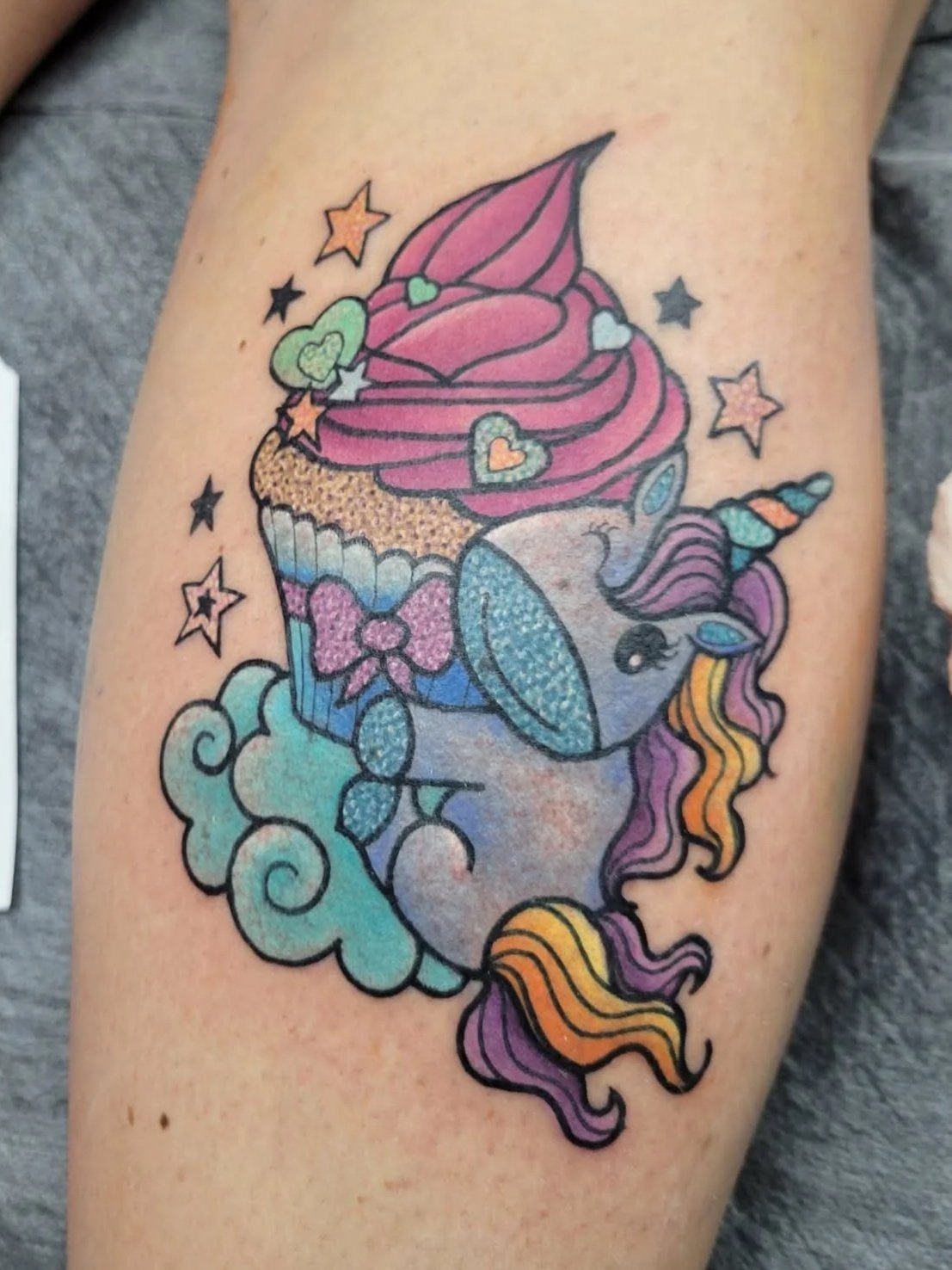 Colorful tattoo of a unicorn with closed eyes, a rainbow-colored mane, and a horn, surrounded by stars and clouds, with a cupcake design featuring pink frosting and various decorative elements.