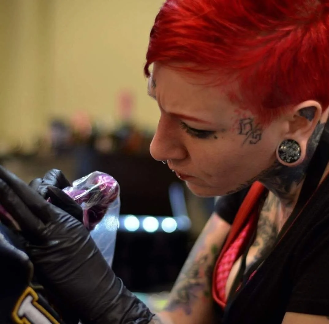 A woman with red hair, tattoos, and gauged ears is holding a small plastic bag with a dark-colored item. She is wearing black gloves and appears to be examining the item closely.