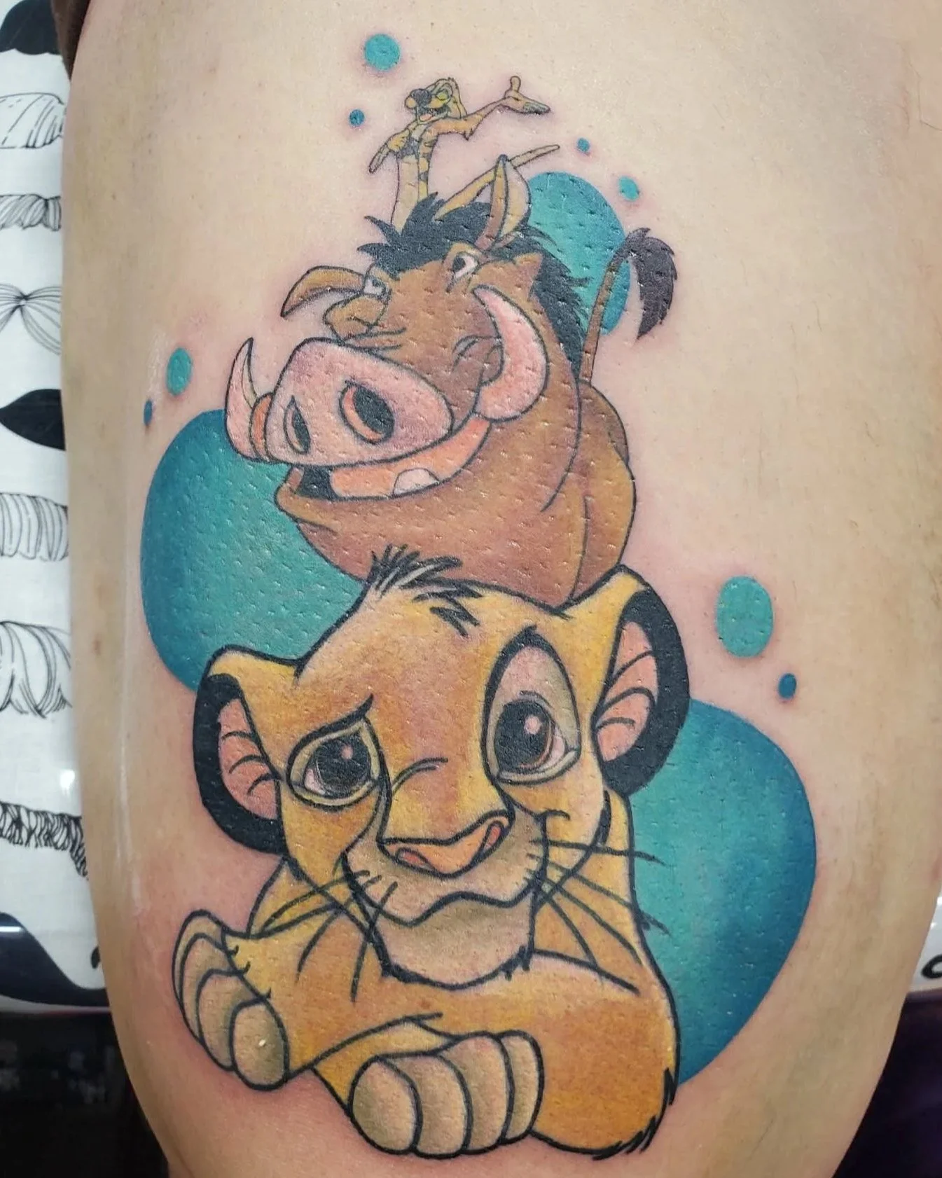 Color tattoo of characters from The Lion King with a lion cub at the bottom, a warthog in the middle, and a meerkat at the top, surrounded by blue circles.