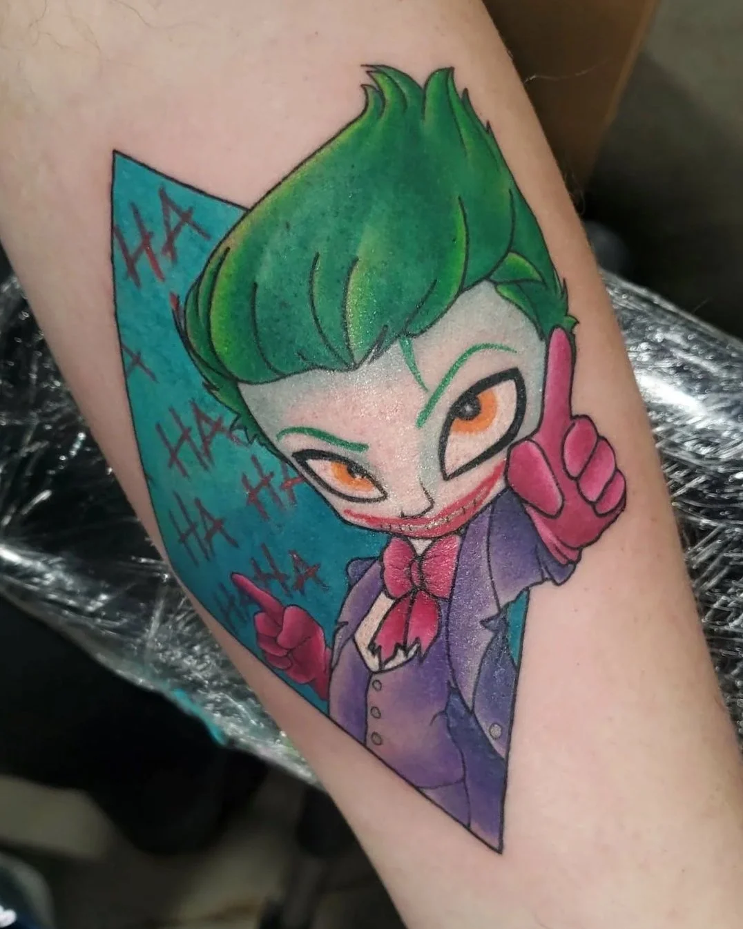 Colorful tattoo of DC Joker cartoon character with green hair, orange eyes, a mischievous expression, and a purple outfit, giving a thumbs-up and sticking out the tongue, with blue background and red 'HA' text.