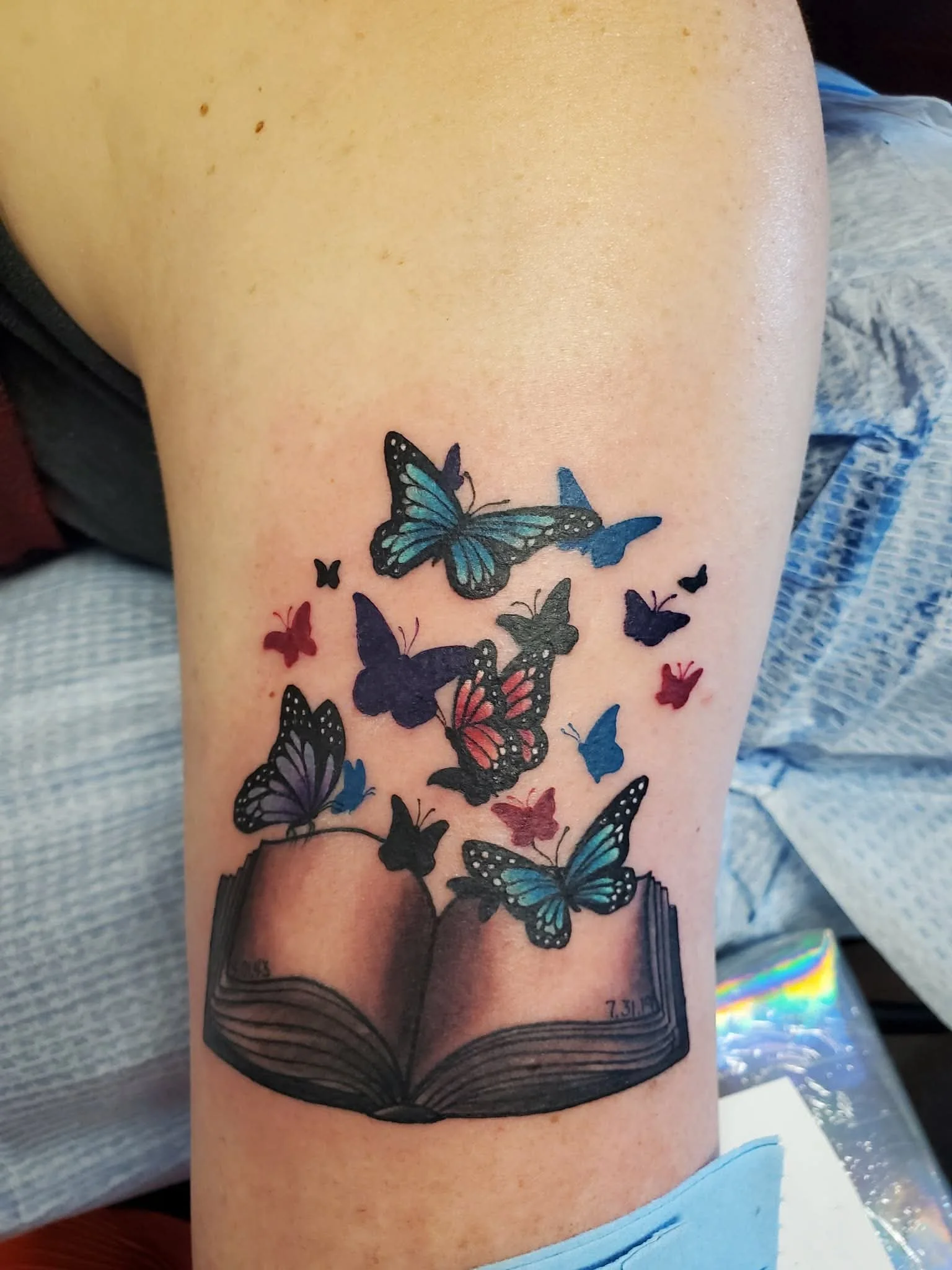 Tattoo of an open book with colorful butterflies emerging from its pages on a person's thigh.