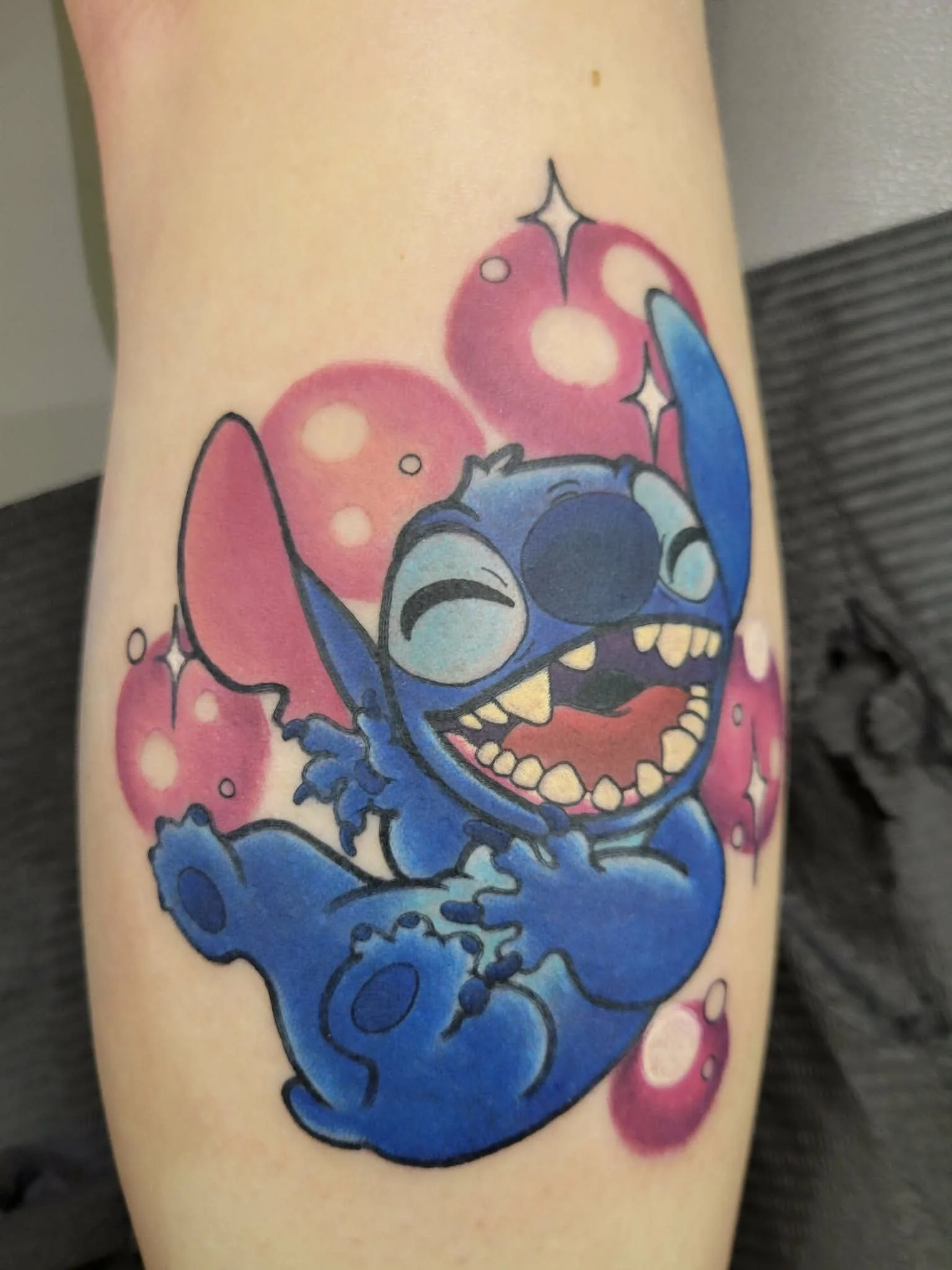 Colorful tattoo of Stitch from Disney's Lilo & Stitch, smiling with big teeth, surrounded by pink bubbles on the skin.
