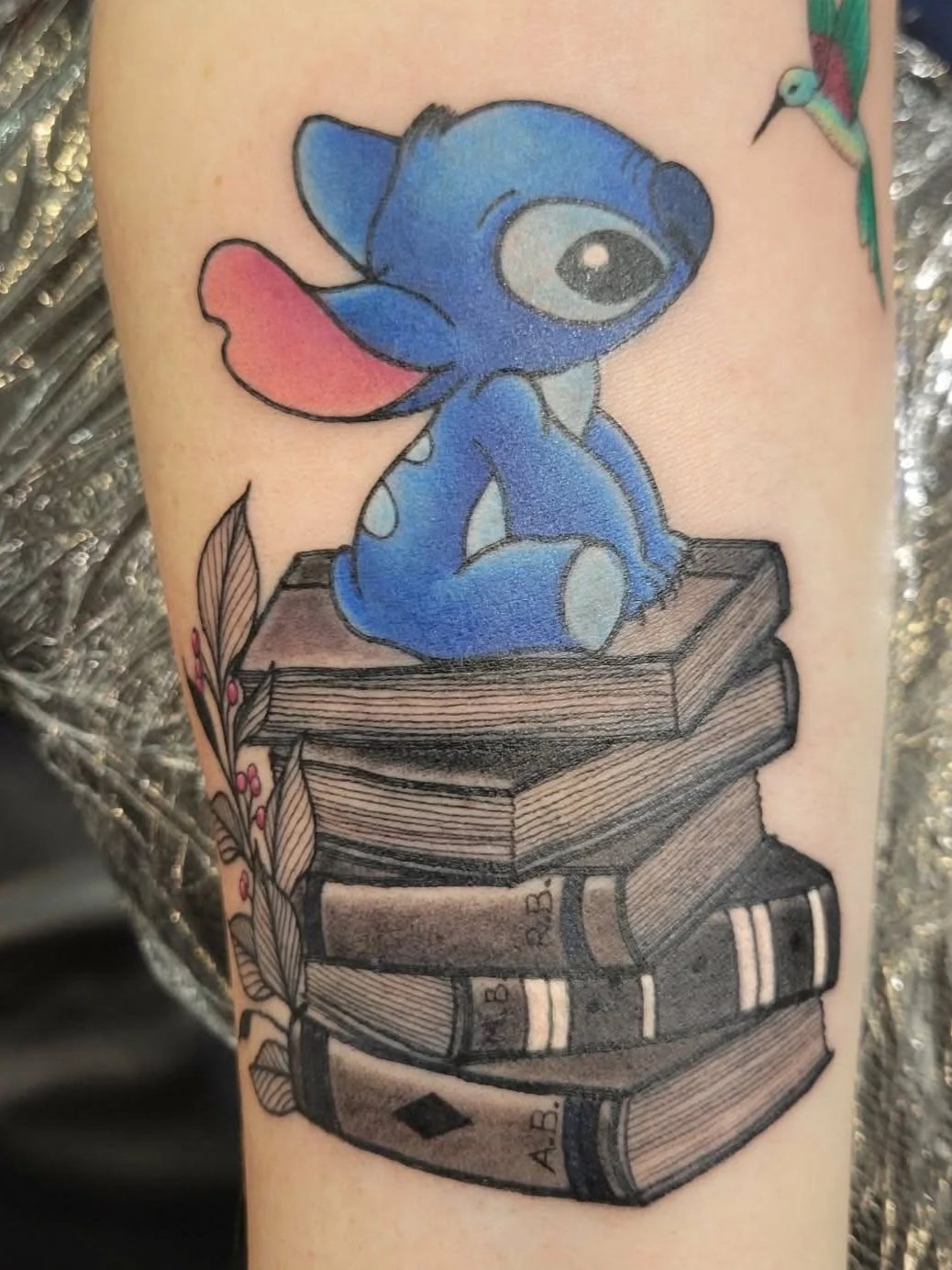 Colorful tattoo of Stitch from Disney's Lilo & Stitch sitting on a stack of books next to a feathered plant and a small flying hummingbird.