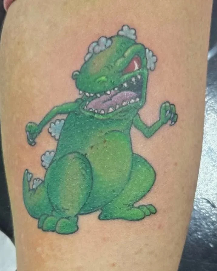 A colorful tattoo of Reptar with a cartoonish appearance, green body, open mouth, and small smoke puffs around its head.