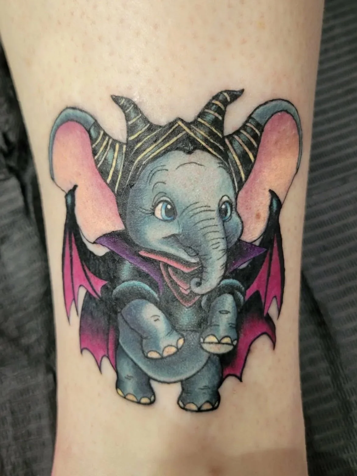 Colorful tattoo of a cute Disney Dumbo wearing Disney Malicifent costume, on person's arm.