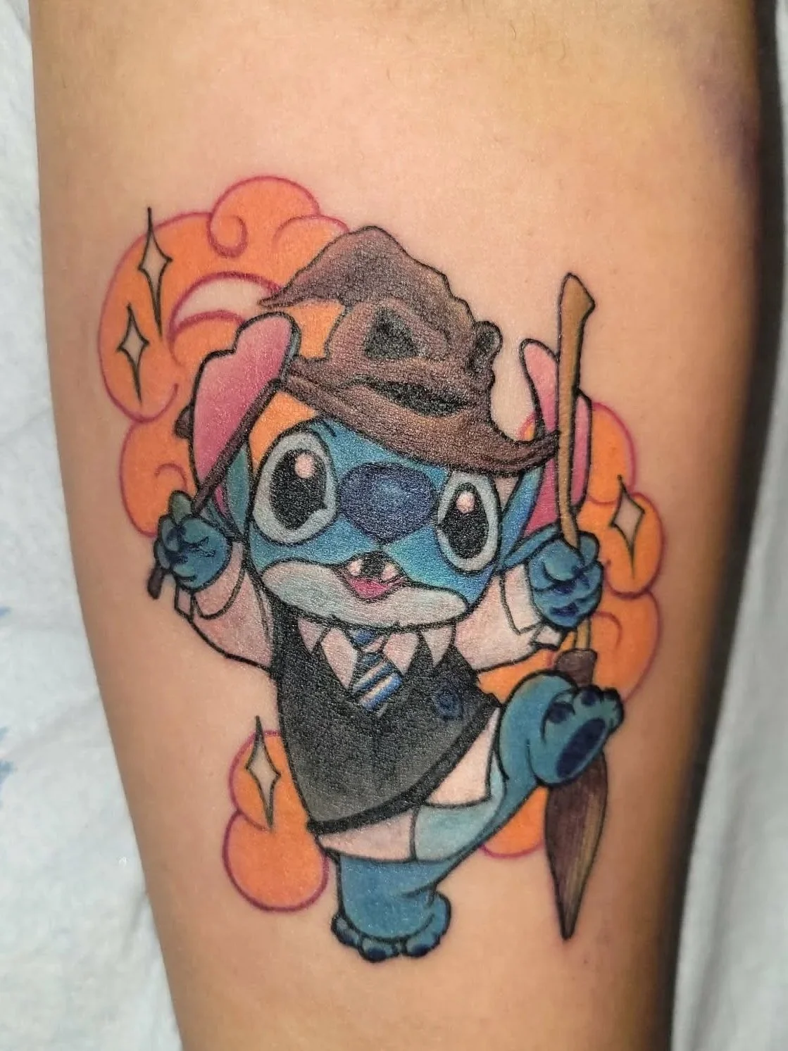 Colorful tattoo of Stitch from Disney's Lilo & Stitch dressed as Harry Potter, with a wizard's robe, glasses, and a hat, holding a broomstick, surrounded by orange sparkles.