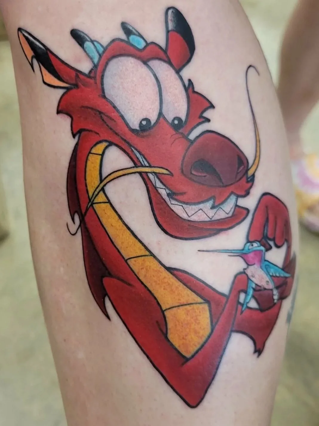 Tattoo of Disney's animated character, Mushu, holding a small blue and pink hummingbird.