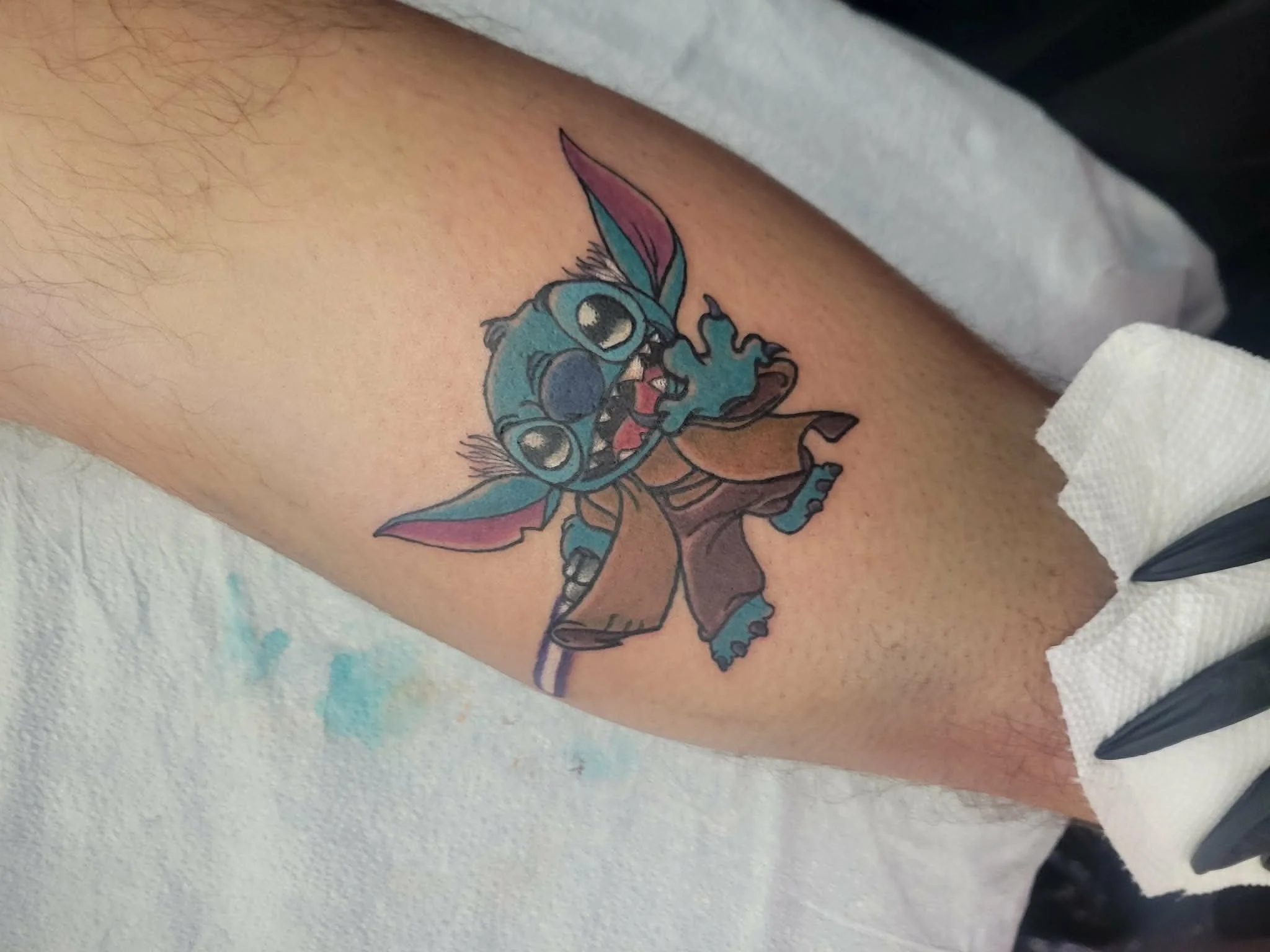Colorful tattoo of Stitch from Disney's Lilo & Stitch on person's arm, showing Stitch with big eyes, tongue out, and wearing brown shorts.