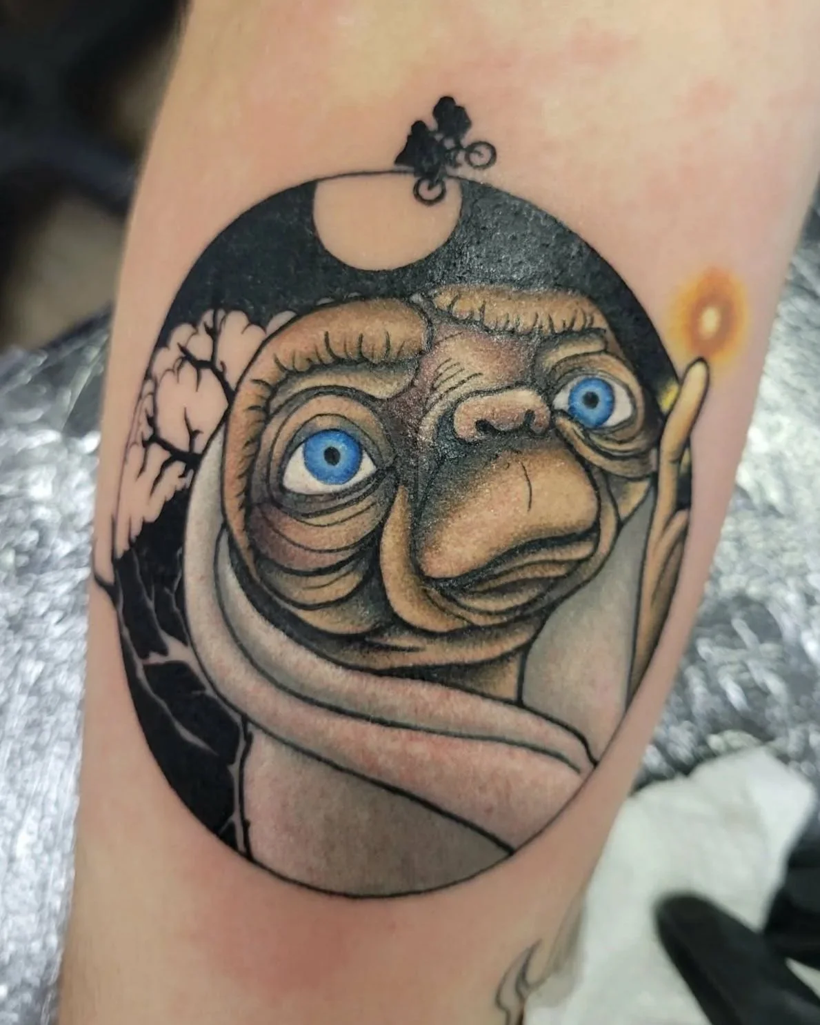 Tattoo of movie character E.T. with blue eyes, inside a circular frame with a moon and a jumping cyclist silhouette at the top.