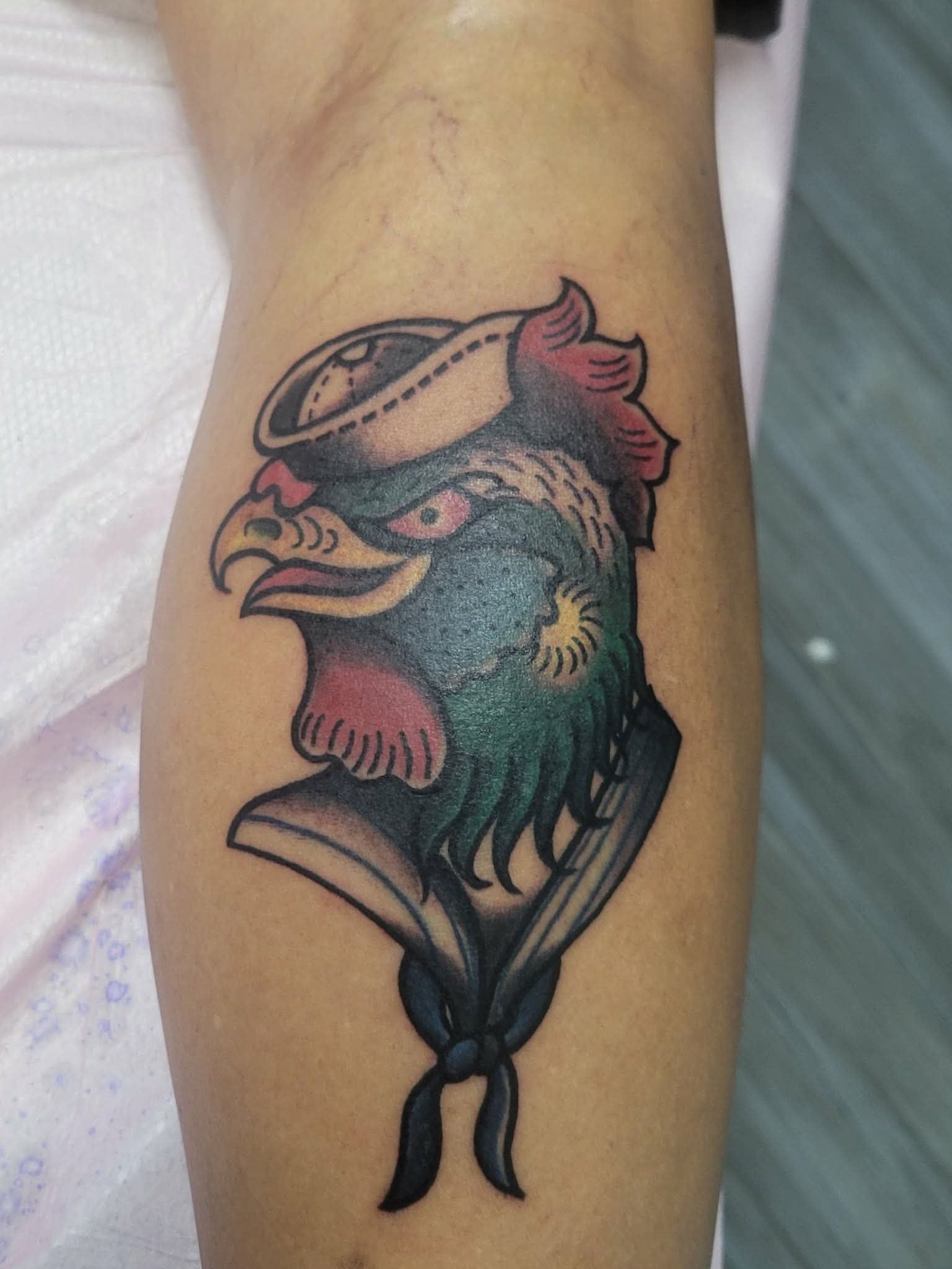 Tattoo of a colorful rooster head in a traditional style with a sailor hat, on person's arm.