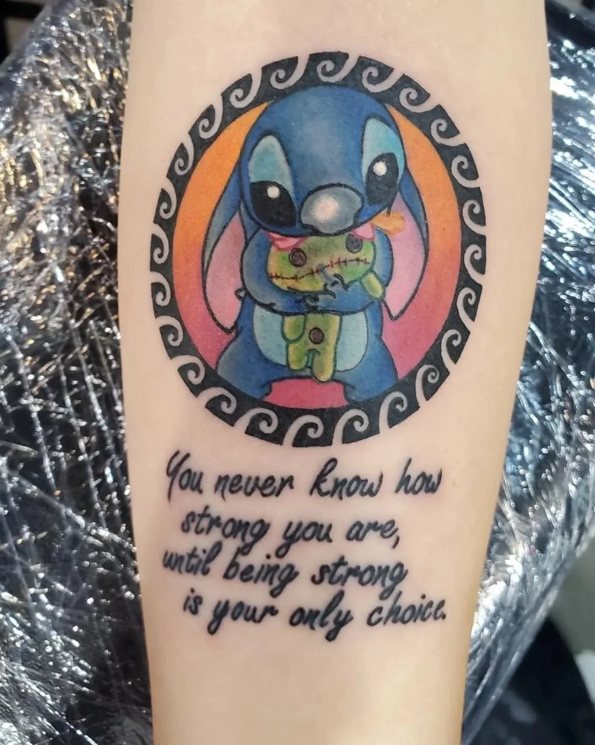A colorful tattoo of Stitch from Lilo & Stitch holding a stuffed toy, with a quote that reads, "You never know how strong you are, until being strong is your only choice."