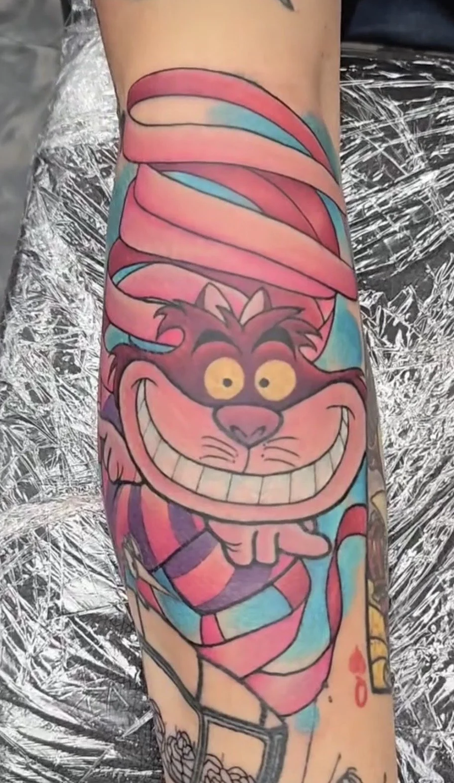 Colorful tattoo of the Cheshire Cat from Alice in Wonderland on a person's arm, with pink, purple, and blue hues and a wide grin.