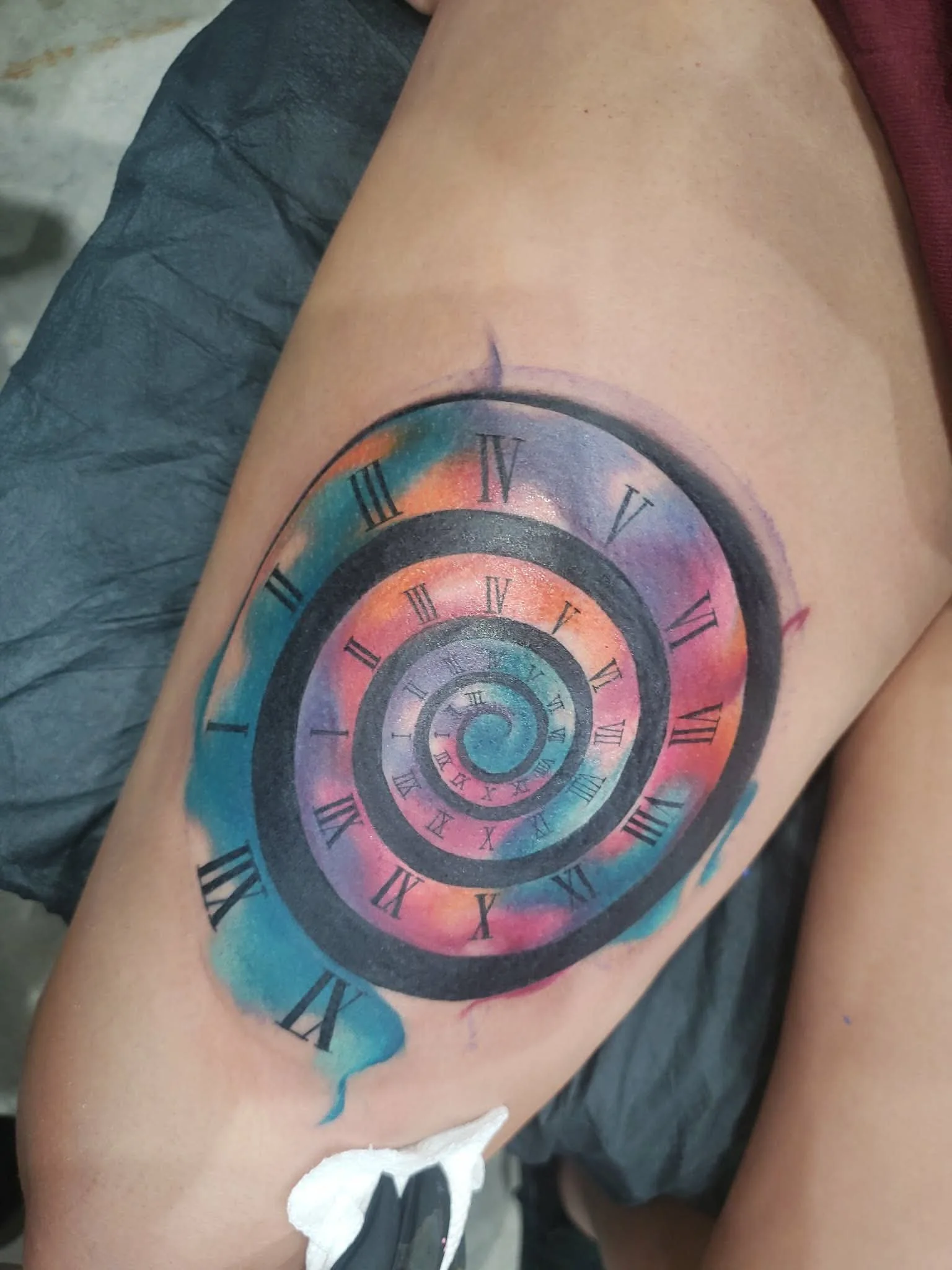 Colorful tattoo on thigh featuring a spiral galaxy with finger-painted watercolor effects and overlaid black Roman numeral clock face.