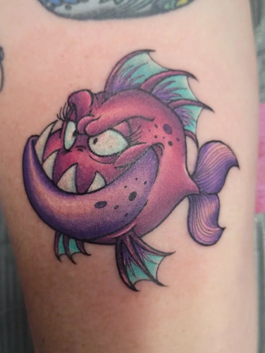 Colorful tattoo of a cartoonish anglerfish with purple, pink, and green colors, showing sharp teeth.