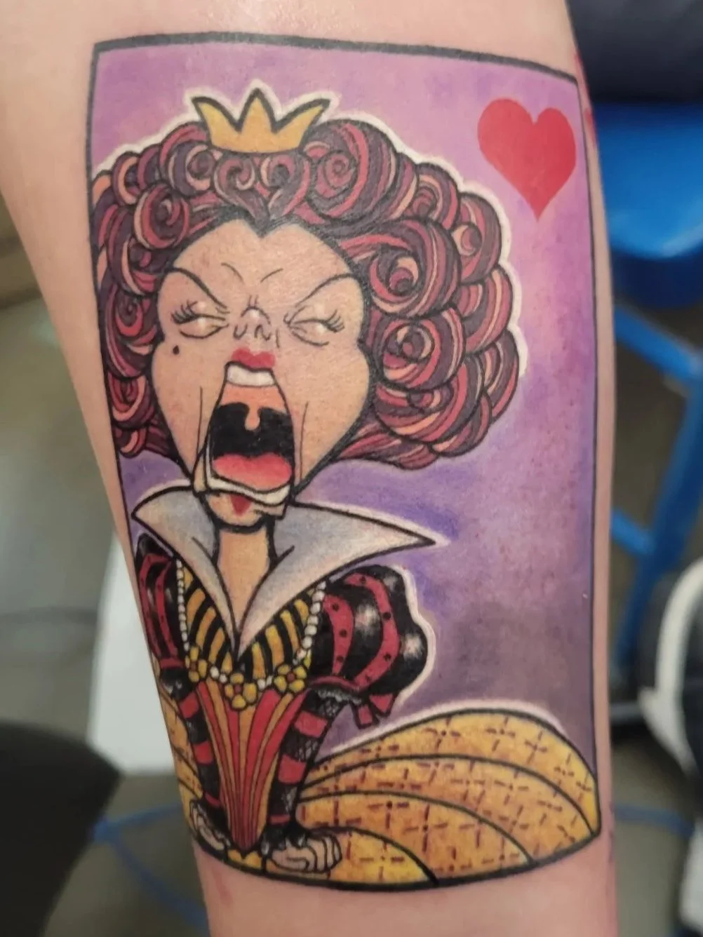Tattoo of a cartoon queen with curly hair, a small crown, a red heart, and a yellow-patterned hat in the background.