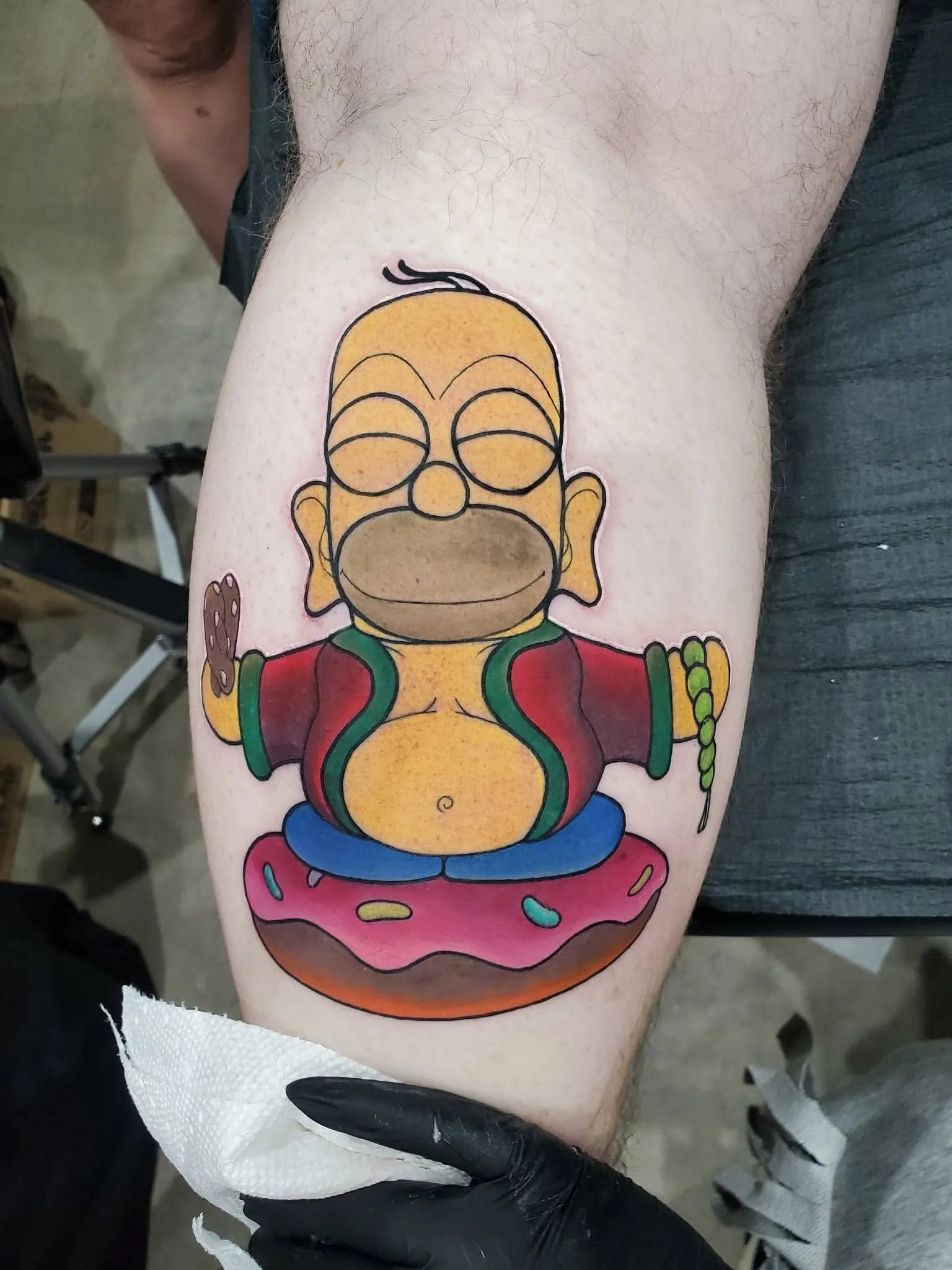 Colorful cartoon tattoo of Simpson’s Homer character with glasses, a red and green jacket, and sitting on a pink donut with sprinkles.